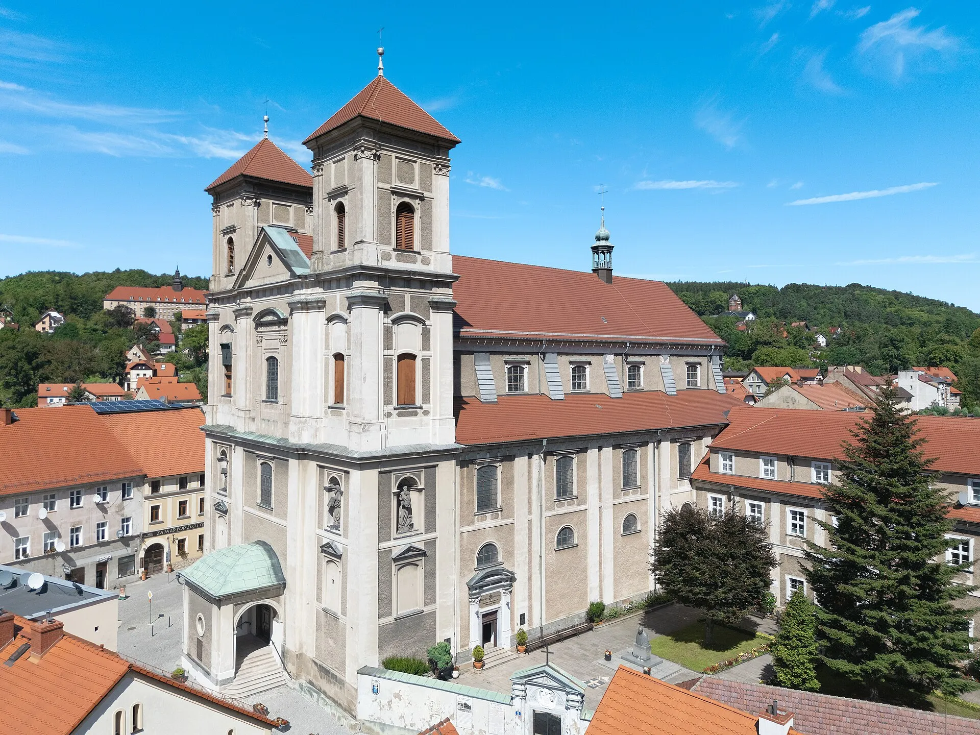 Bazylika Nawiedzenia Najświętszej Maryi Panny w Bardzie z lotu ptaka: dwuwieżowa fasada i nawa z czerwonymi dachami wśród zabudowy miasta i zalesionych wzgórz