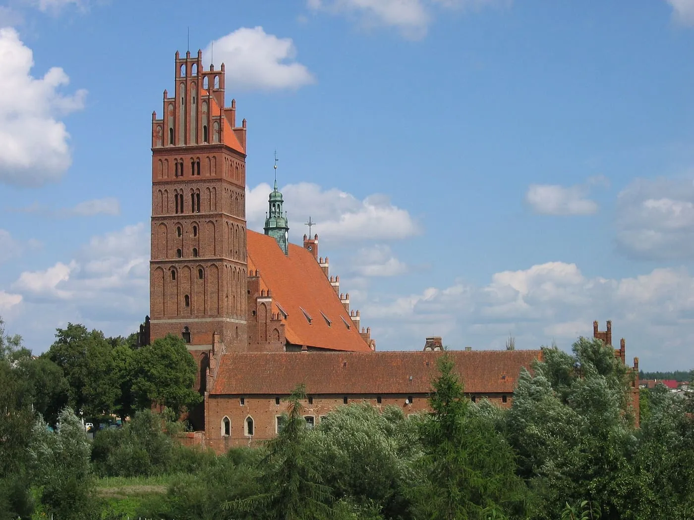 Gotycka bazylika kolegiacka w Dobrym Mieście z wysoką ceglaną wieżą zwieńczoną schodkowym szczytem, otoczona zielenią