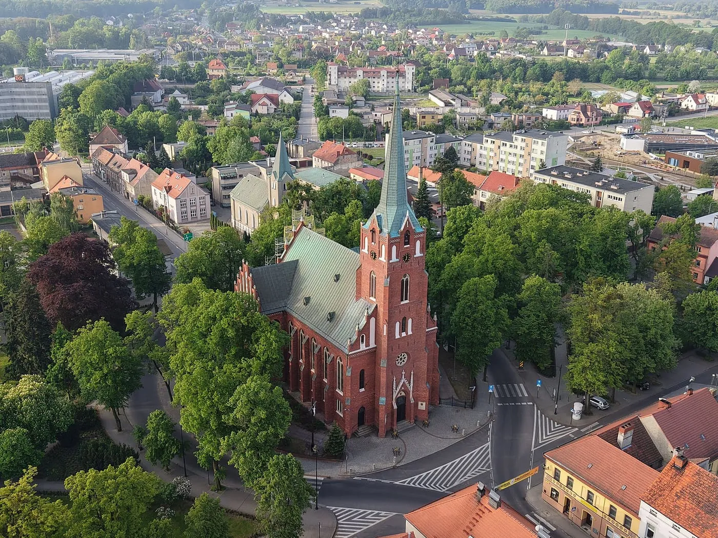 Widok z lotu ptaka na ceglaną bazylikę z wysoką, zwieńczoną iglicą wieżą, otoczoną zieleńcem i uliczkami z kolorowymi kamienicami, w słoneczny dzień