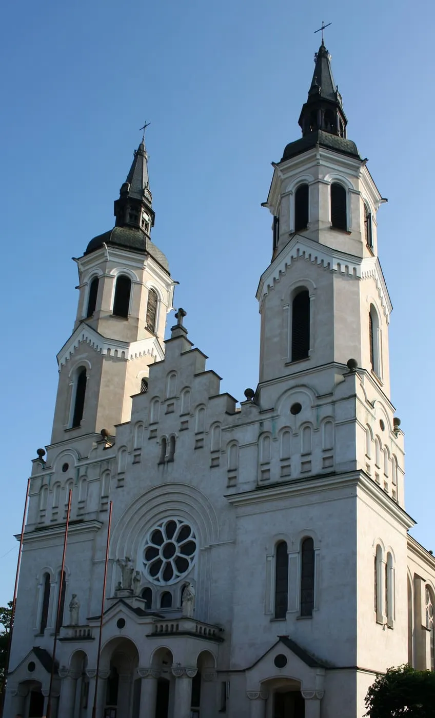 Eklektyczna, kremowa fasada bazyliki w Augustowie z dwiema wieżami, rozetą i figurami świętych