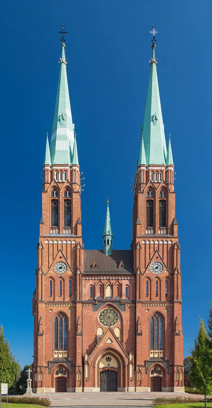 Neogotycka, ceglana fasada bazyliki św. Antoniego w Rybniku z dwiema wysokimi wieżami zwieńczonymi zielonymi, miedzianymi iglicami, rozetą i trzema portalami, na tle granatowego nieba