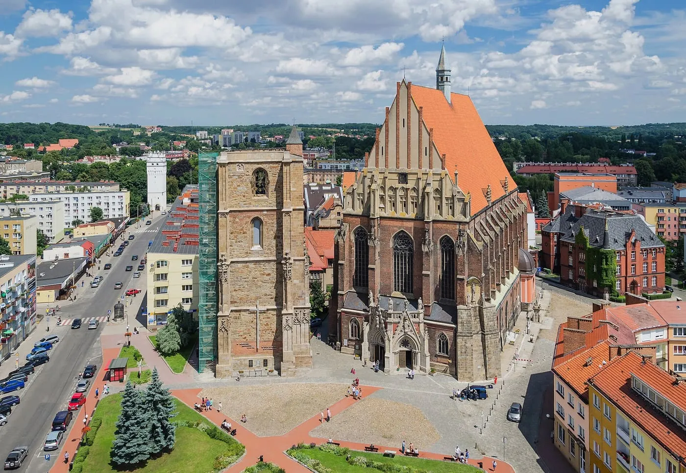 Widok z góry na gotycką bazylikę z pomarańczowym dachem i schodkowym szczytem oraz osobną kamienną dzwonnicę z rusztowaniem, wokół plac i ulice miasta