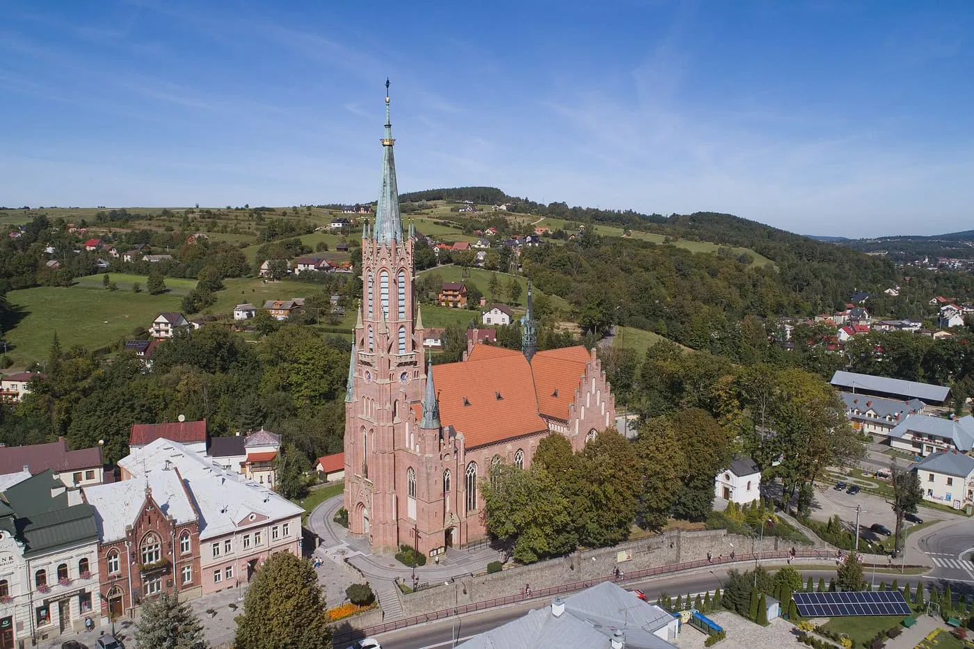 Widok z góry na różowy, neogotycki kościół z bardzo wysoką, smukłą wieżą, wznoszący się nad miasteczkiem otoczonym zielonymi wzgórzami, w słoneczny dzień