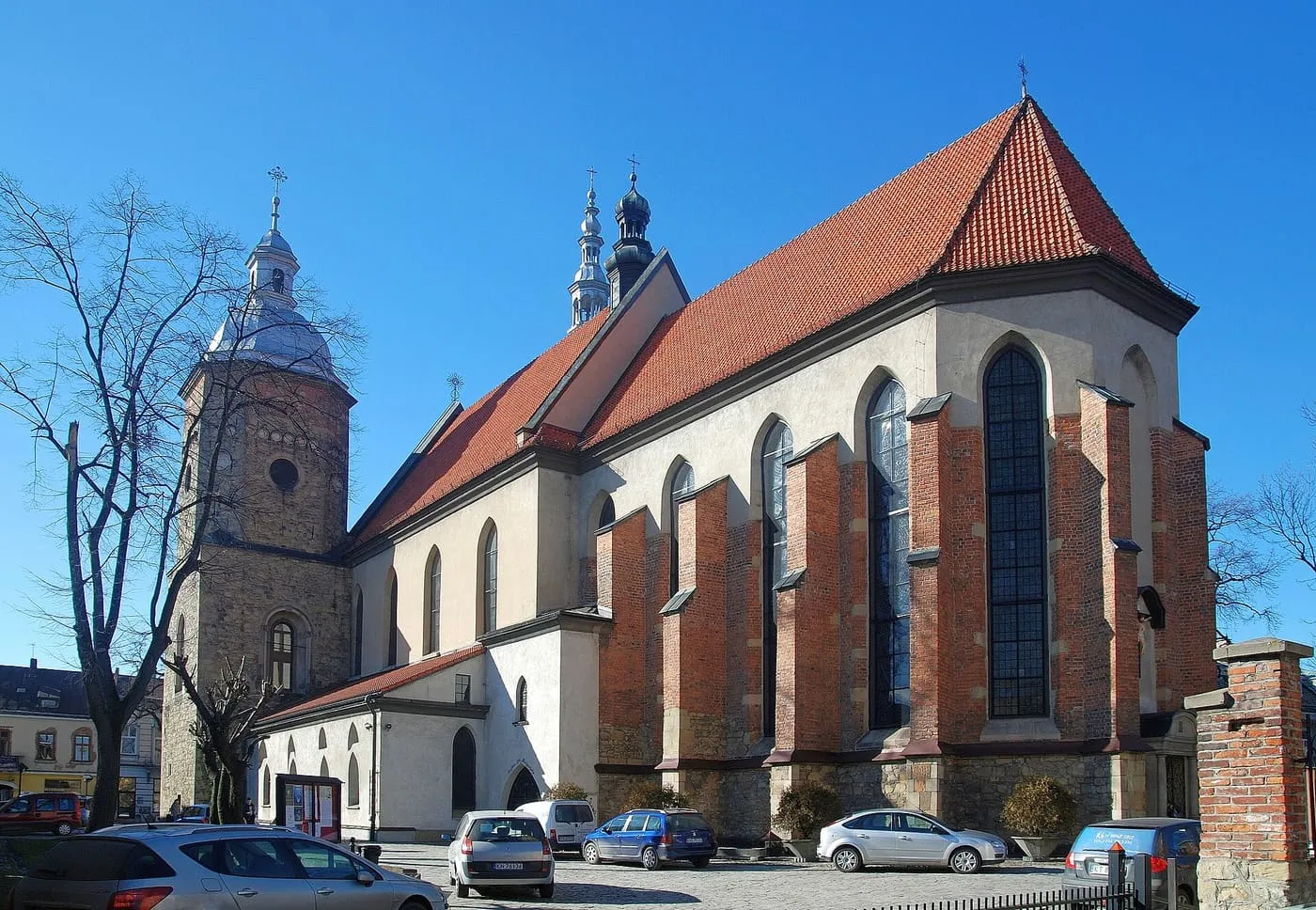 Gotycka bazylika św. Małgorzaty w Nowym Sączu widziana od strony prezbiterium, z ceglanymi szkarpami, czerwonym dachem i kamienną wieżą zwieńczoną białą, barokową latarnią