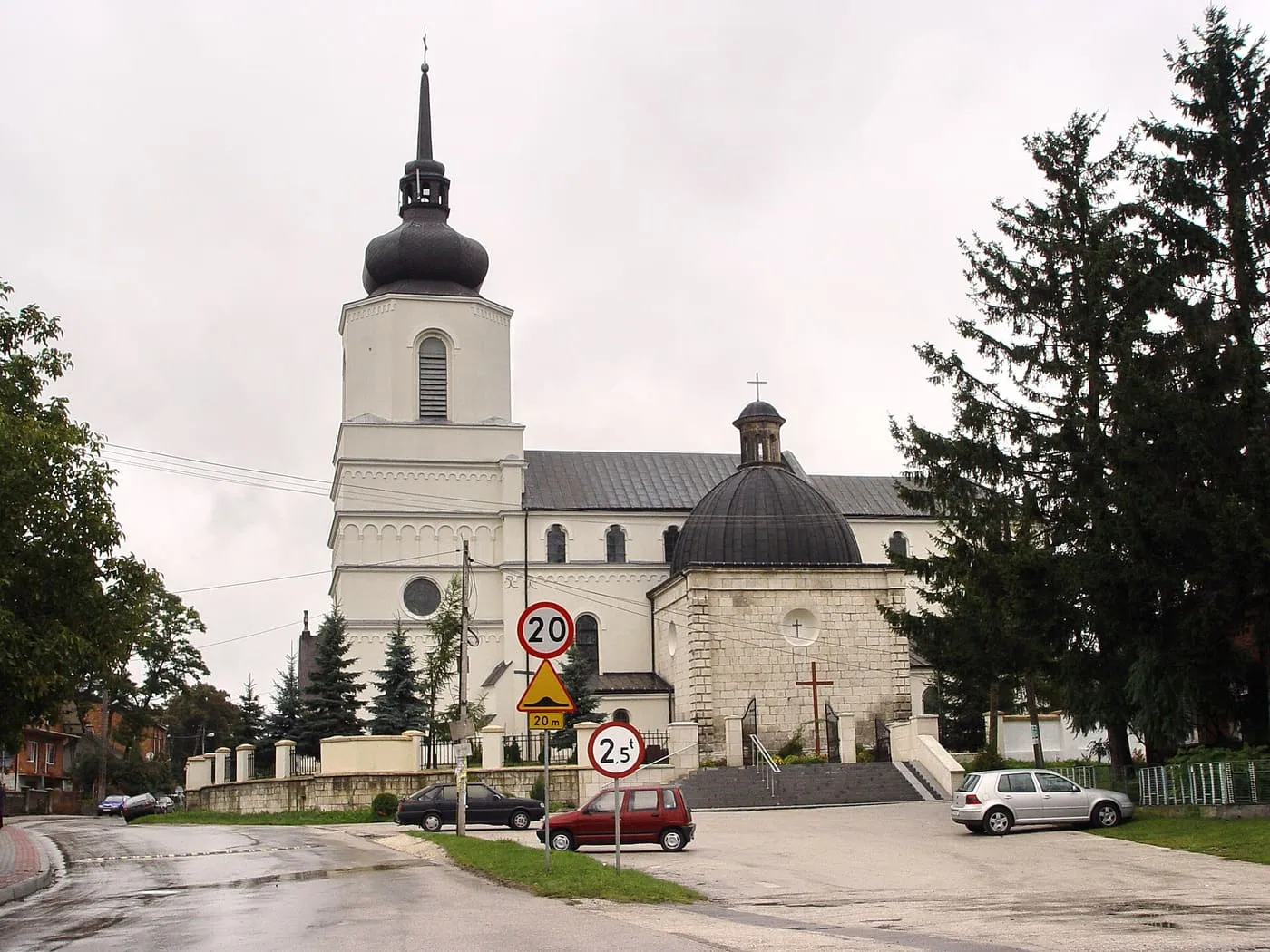Biała wieża z ciemnym, cebulastym hełmem i kamienna kopuła kaplicy przy bazylice, widziane od strony jezdni wśród świerków, w pochmurny dzień