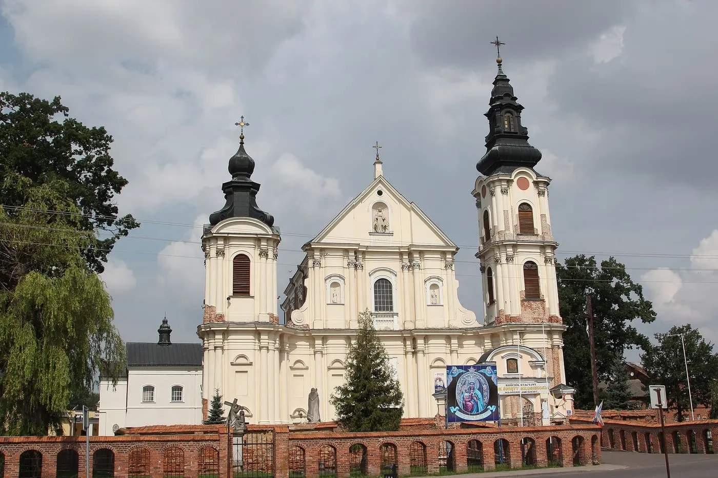 Kremowa, dwuwieżowa fasada barokowej bazyliki z ciemnymi hełmami wież i falującym szczytem, przed którą biegnie niski ceglany mur z arkadami