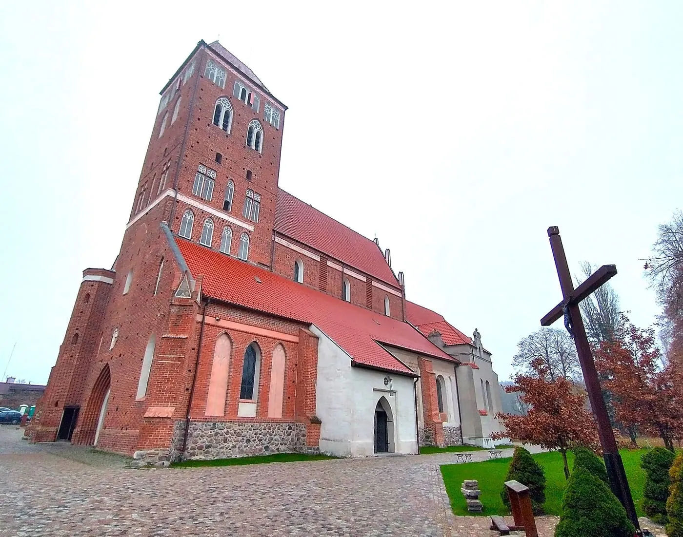 Gotycka, ceglana bazylika św. Tomasza w Nowym Mieście Lubawskim z masywną czworoboczną wieżą, czerwonymi dachami i bielonymi przybudówkami na kamiennym cokole, obok duży drewniany krzyż
