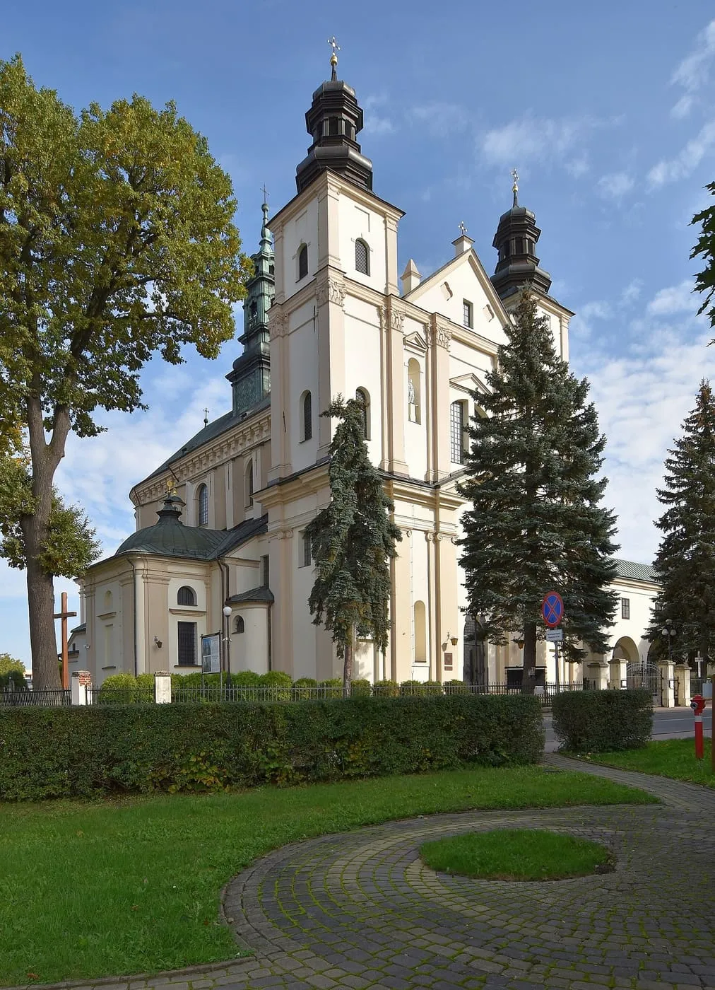 Barokowa, kremowa fasada bazyliki w Brzozowie z dwiema wieżami zwieńczonymi ciemnymi, cebulastymi hełmami, wśród świerków