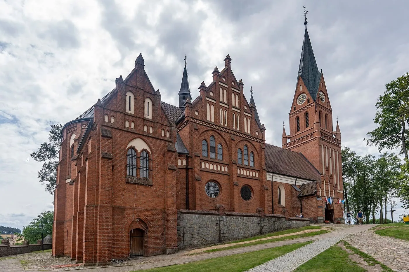 Ceglana, neogotycka bazylika w Gietrzwałdzie ze schodkowymi szczytami i wysoką wieżą z zegarem, z brukowaną drogą prowadzącą do wejścia pod zachmurzonym niebem