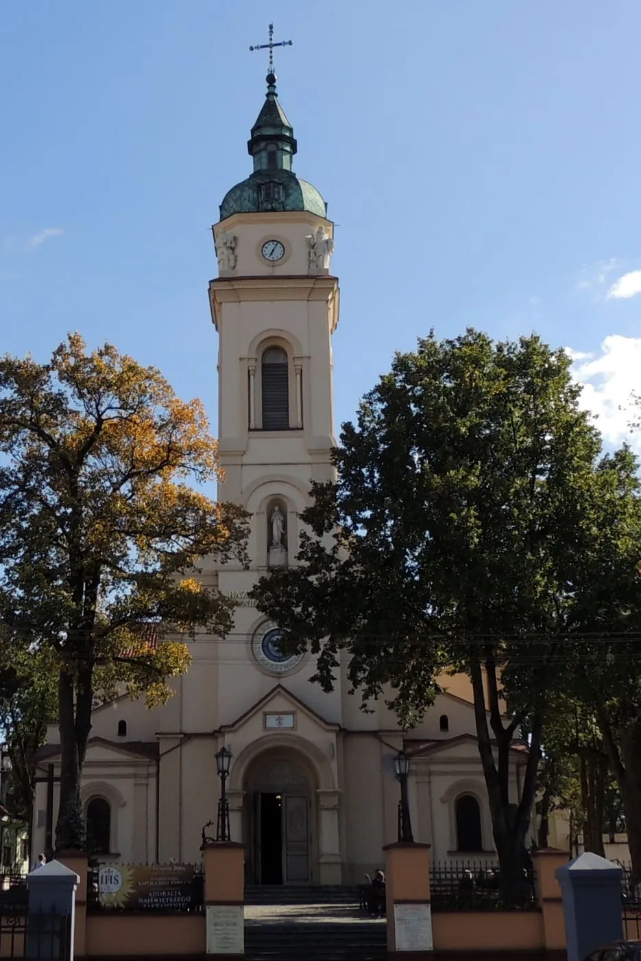 Kremowa bazylika w Zduńskiej Woli z wieżą zwieńczoną zieloną, miedzianą kopułą i latarnią z krzyżem, widziana zza jesiennych drzew
