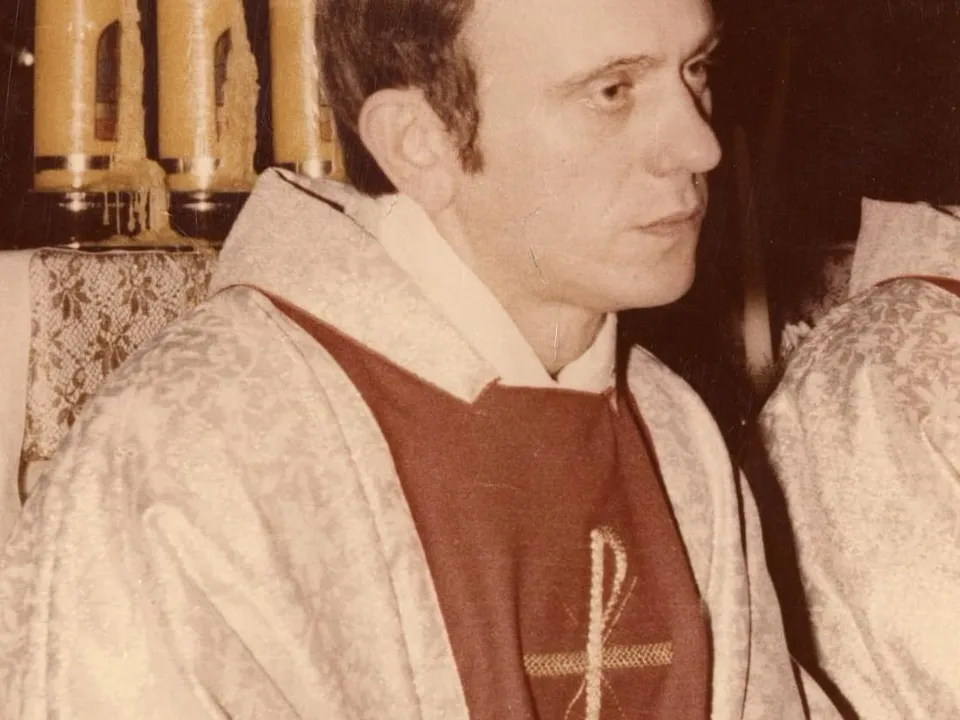Fotografia księdza Jerzego Popiełuszki w szatach liturgicznych: kapłan siedzi w profilu, w jasnej ornatowej szacie z ciemnoczerwoną wstawką ozdobioną chrystogramem i kłosami, a za nim płoną świece przy ołtarzu