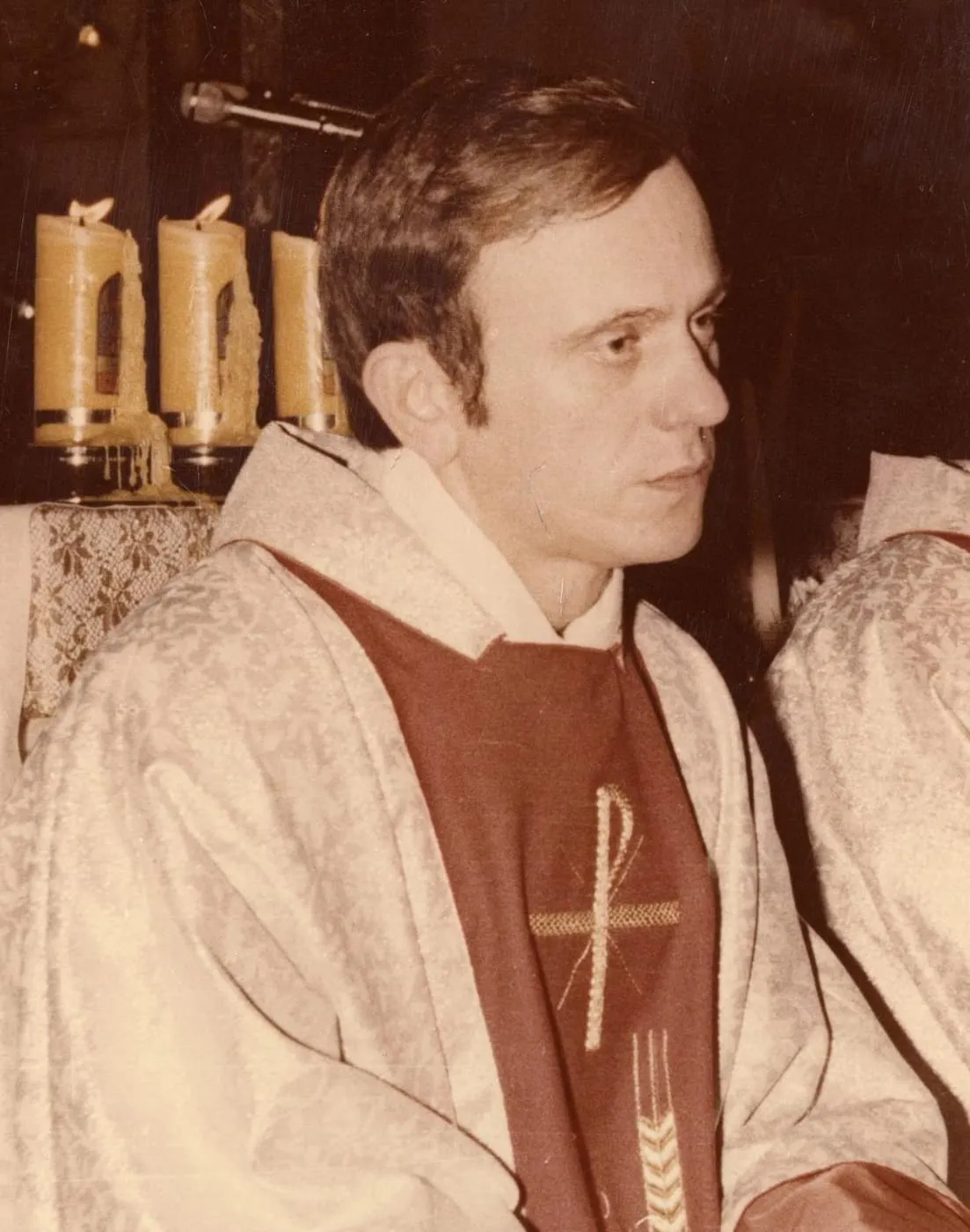 Fotografia księdza Jerzego Popiełuszki w szatach liturgicznych: kapłan siedzi w profilu, w jasnej ornatowej szacie z ciemnoczerwoną wstawką ozdobioną chrystogramem i kłosami, a za nim płoną świece przy ołtarzu