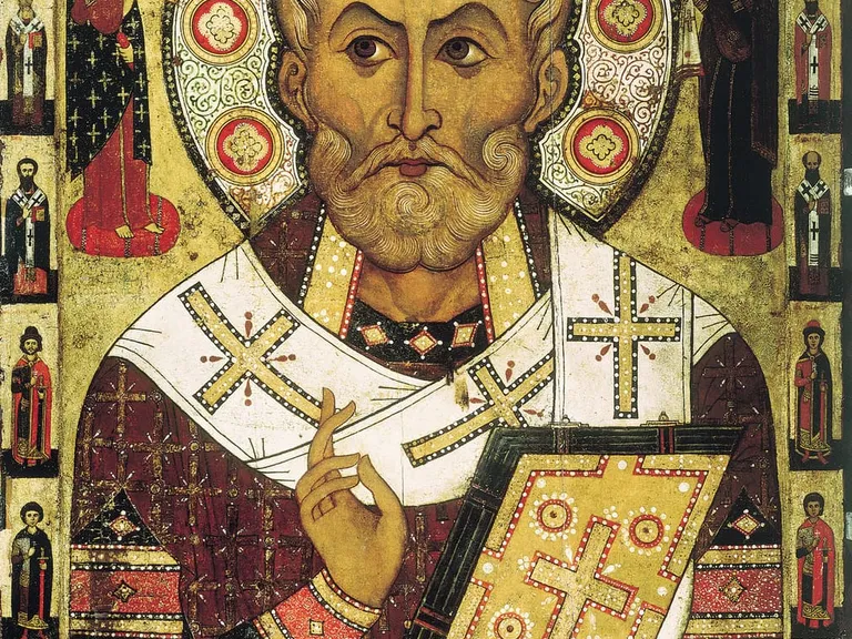 Staroruska ikona św. Mikołaja: biskup z aureolą, w białym omoforionie z krzyżami, z Ewangelią w lewej ręce i prawą uniesioną do błogosławieństwa, obok Chrystus i Maryja, wokół półfigury świętych