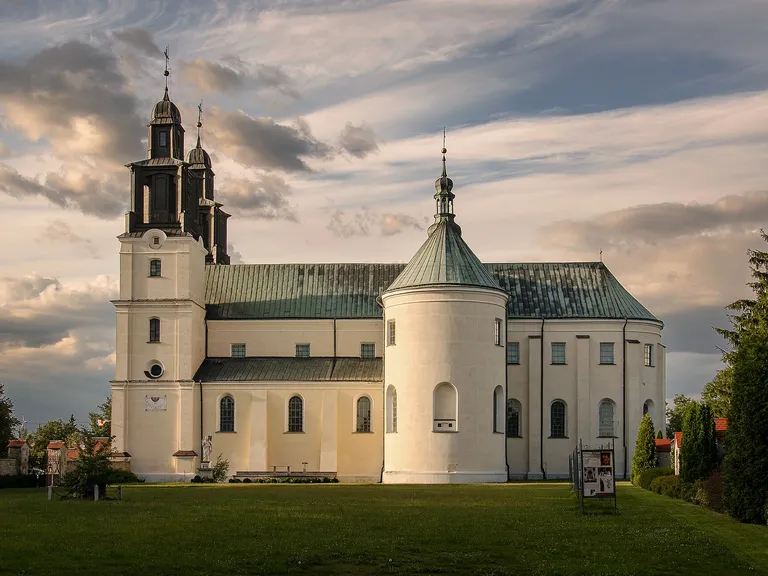Bazylika w Gidlach o zachodzie słońca: jasna bryła z zieloną blaszaną dachówką, dwiema ciemnymi wieżami i okrągłą kaplicą, na tle dramatycznego nieba