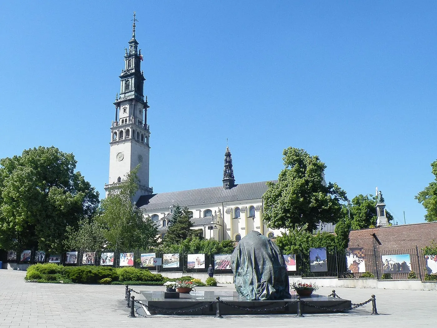 Wieża klasztoru na Jasnej Górze nad placem z drzewami i ogrodzeniem z wystawą zdjęć, pod bezchmurnym niebem w słoneczny dzień