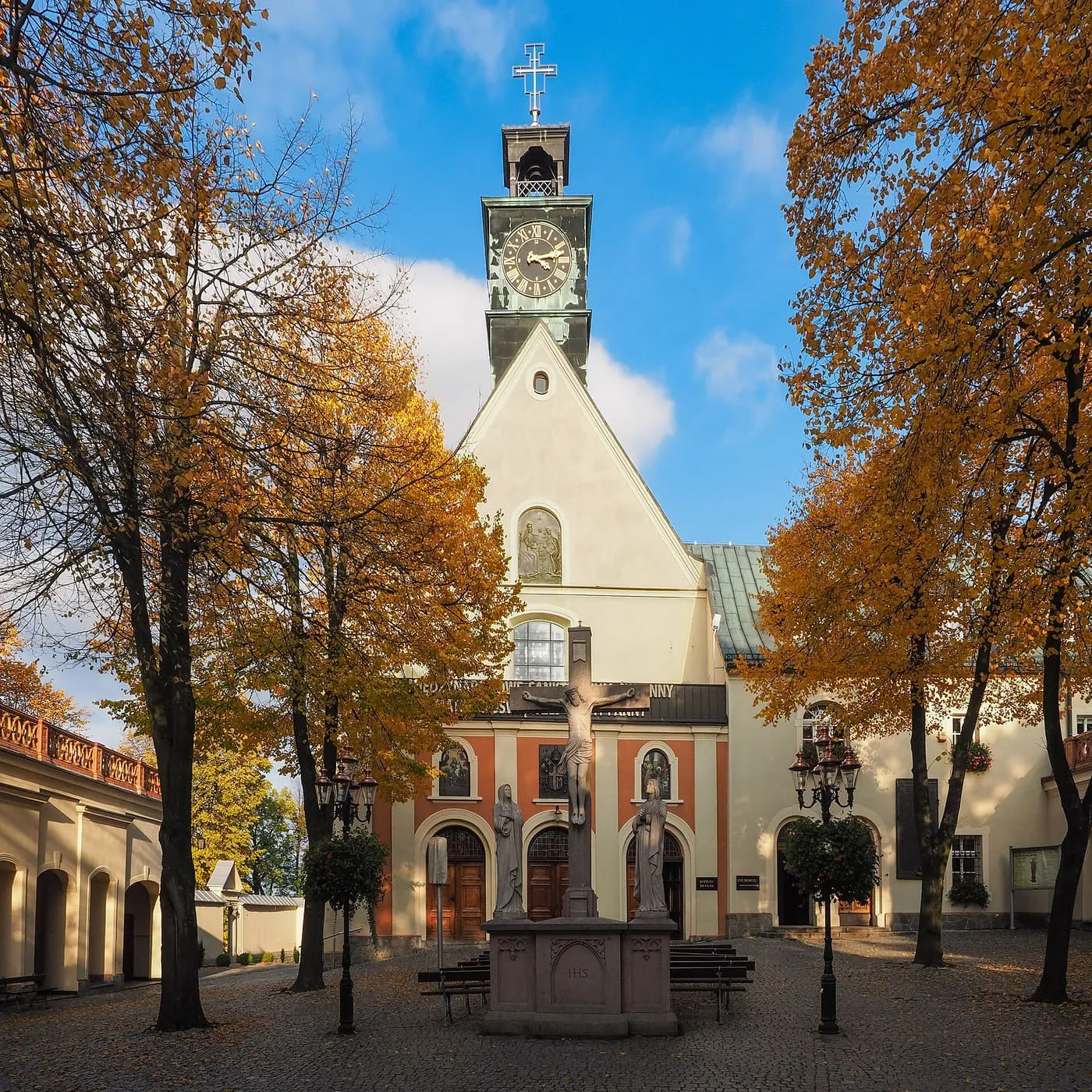 Jesienny dziedziniec przed kościołem sanktuarium: jasna fasada z wieżyczką z zegarem między złocistymi drzewami, przed wejściem krzyż z figurami na cokole