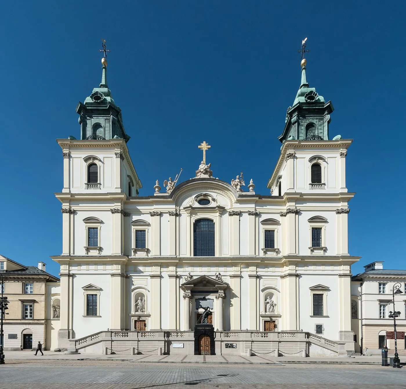 Biała barokowa fasada kościoła Świętego Krzyża w Warszawie z dwiema wieżami o zielonych hełmach, na tle granatowego, bezchmurnego nieba