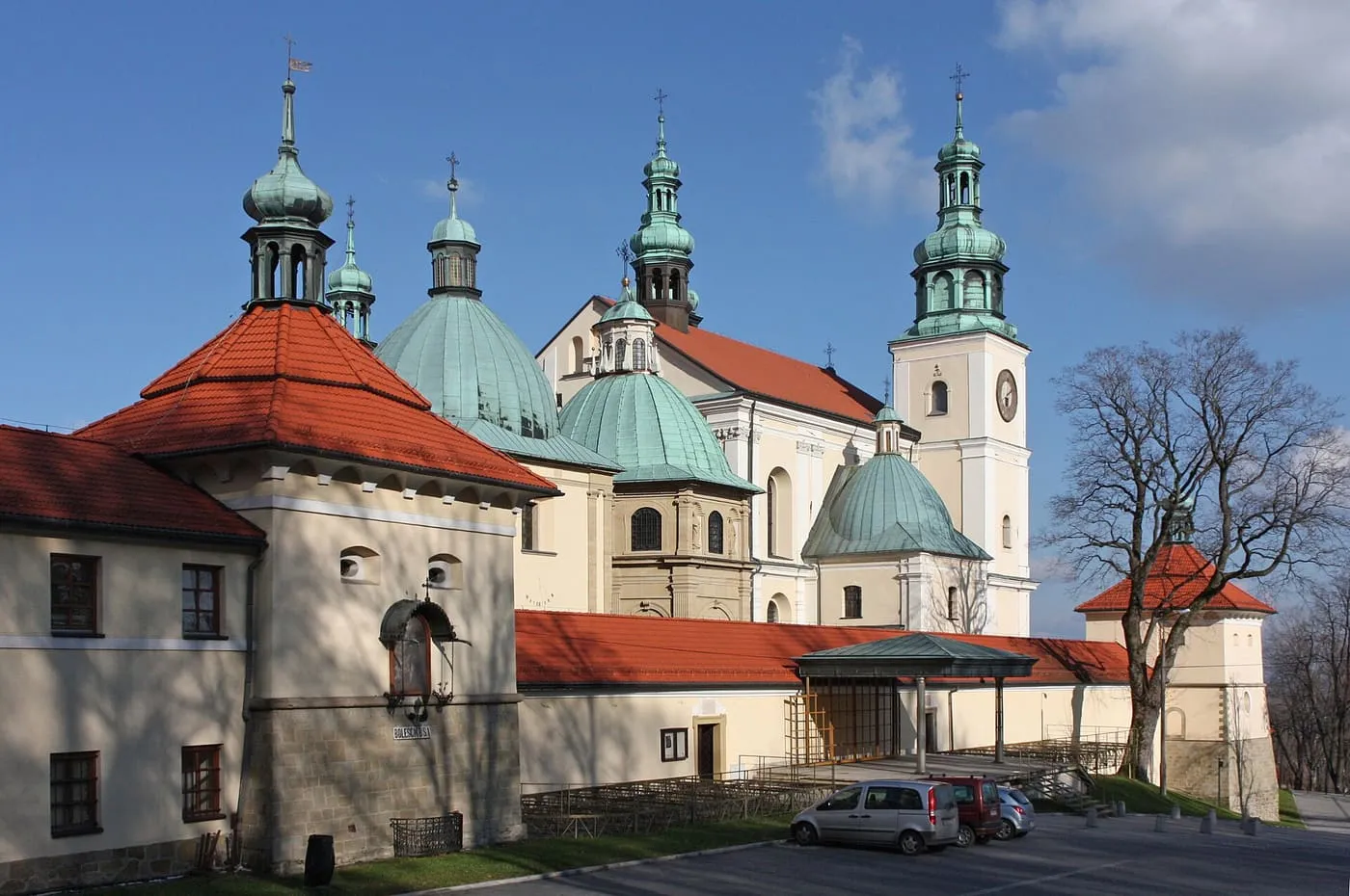 Kompleks bazyliki i klasztoru bernardynów: zielone kopuły z latarniami, pomarańczowe dachy kaplic i klasztoru oraz jasna wieża z zegarem na tle błękitnego nieba