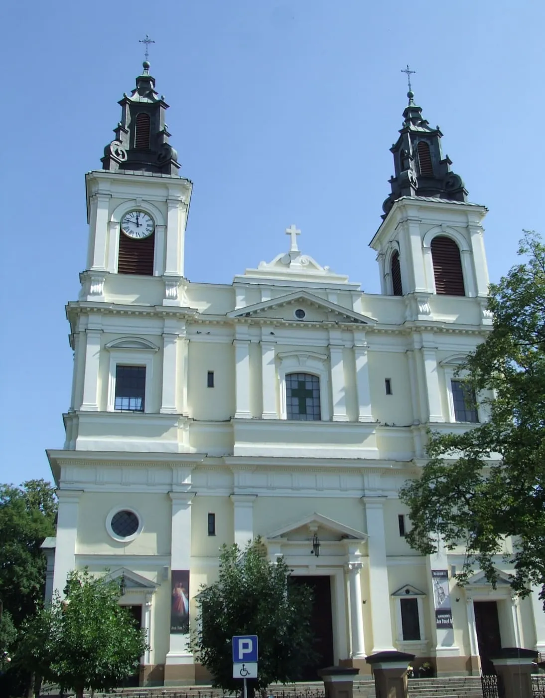 Biała, neobarokowa kolegiata z dwiema wieżami z zegarami, zwieńczonymi ciemnymi hełmami, widziana z ukosa zza drzew, na tle błękitnego nieba