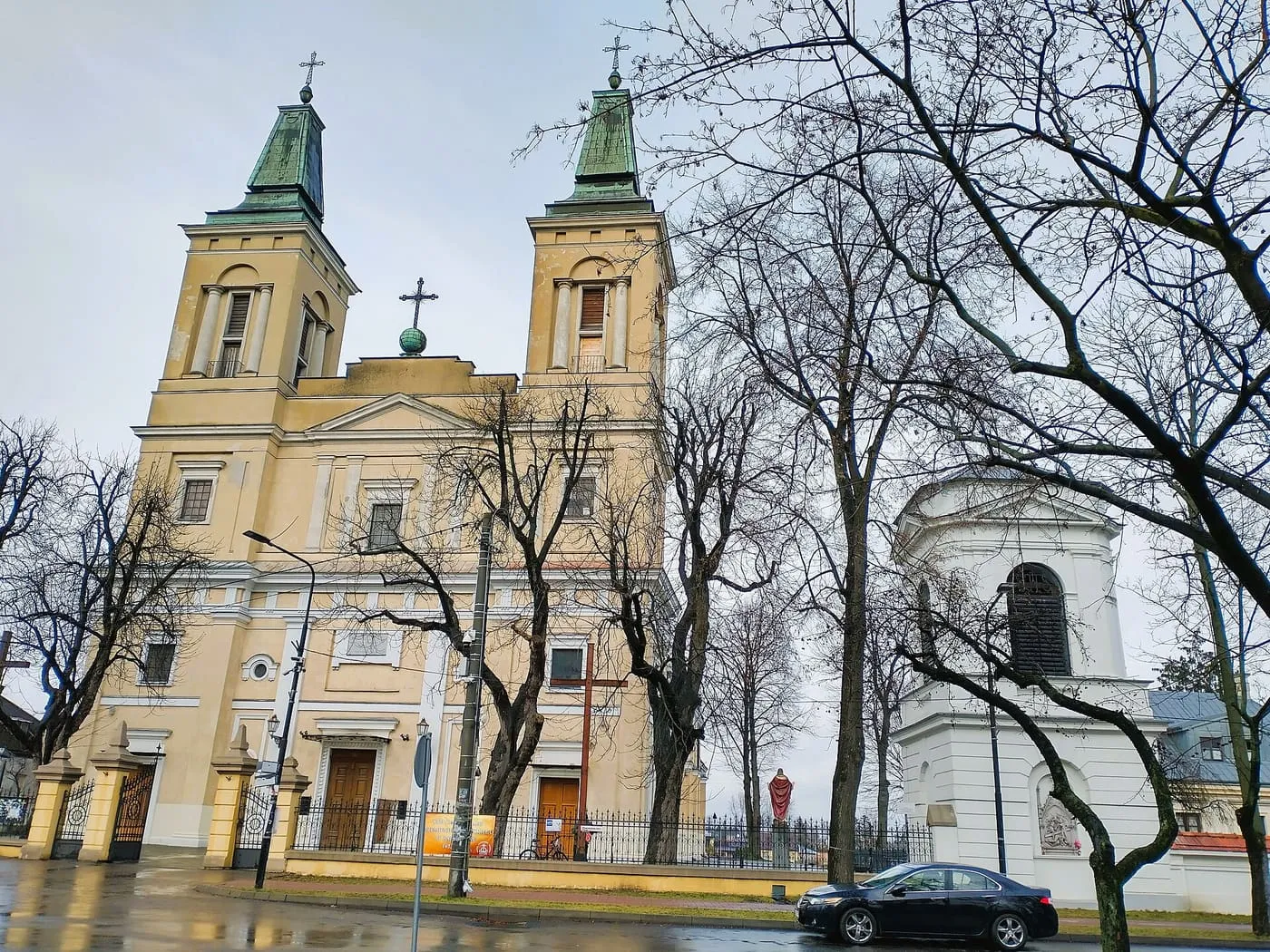 Żółto-kremowa fasada kolegiaty z dwiema wieżami o zielonych hełmach, z boku biała dzwonnica, przed nią ogrodzenie i drzewa