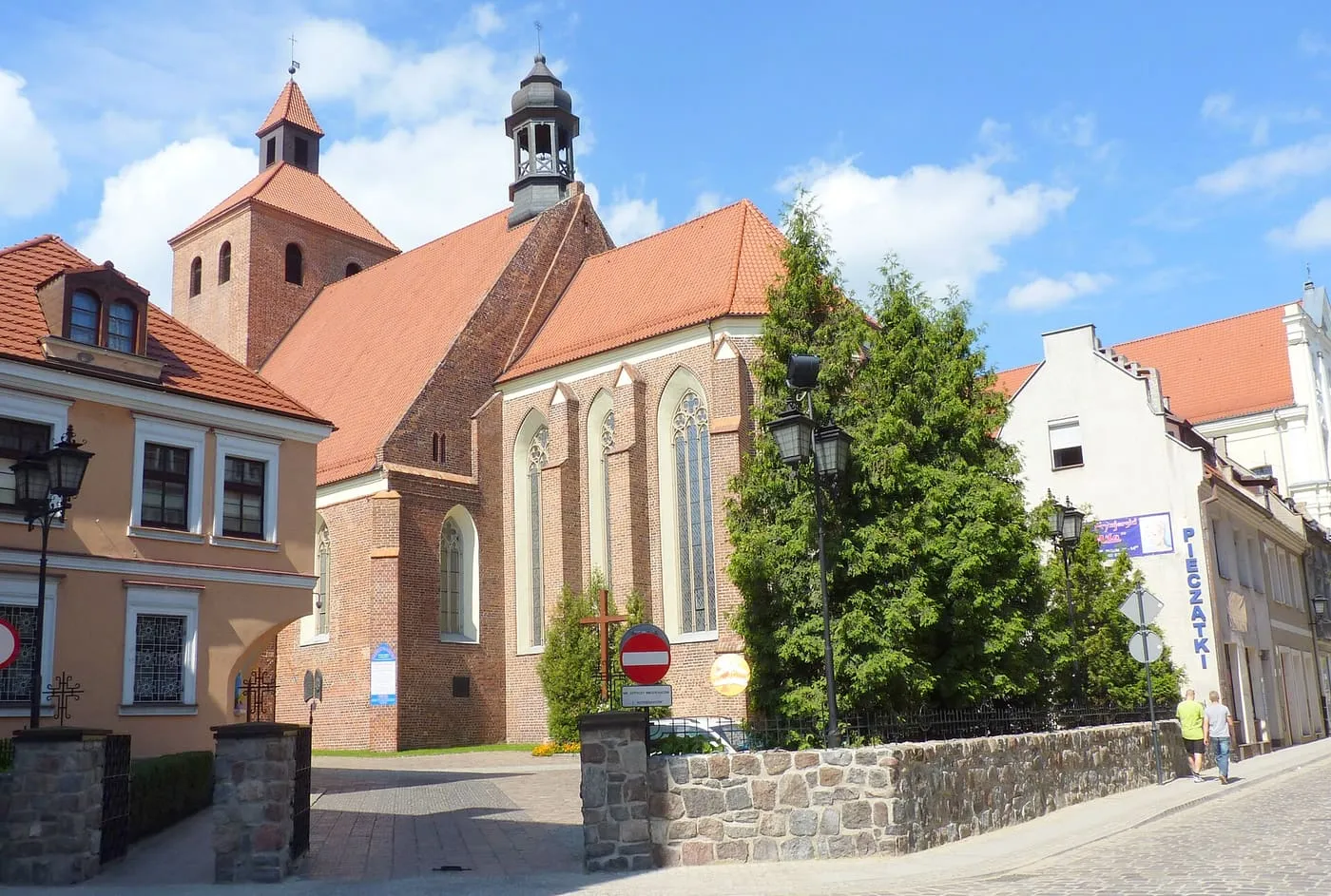 Gotycka, ceglana fara św. Mikołaja w Grudziądzu z wieżą nakrytą czerwonym dachem, ażurową sygnaturką i wysokimi ostrołukowymi oknami, za kamiennym murkiem przy ulicy