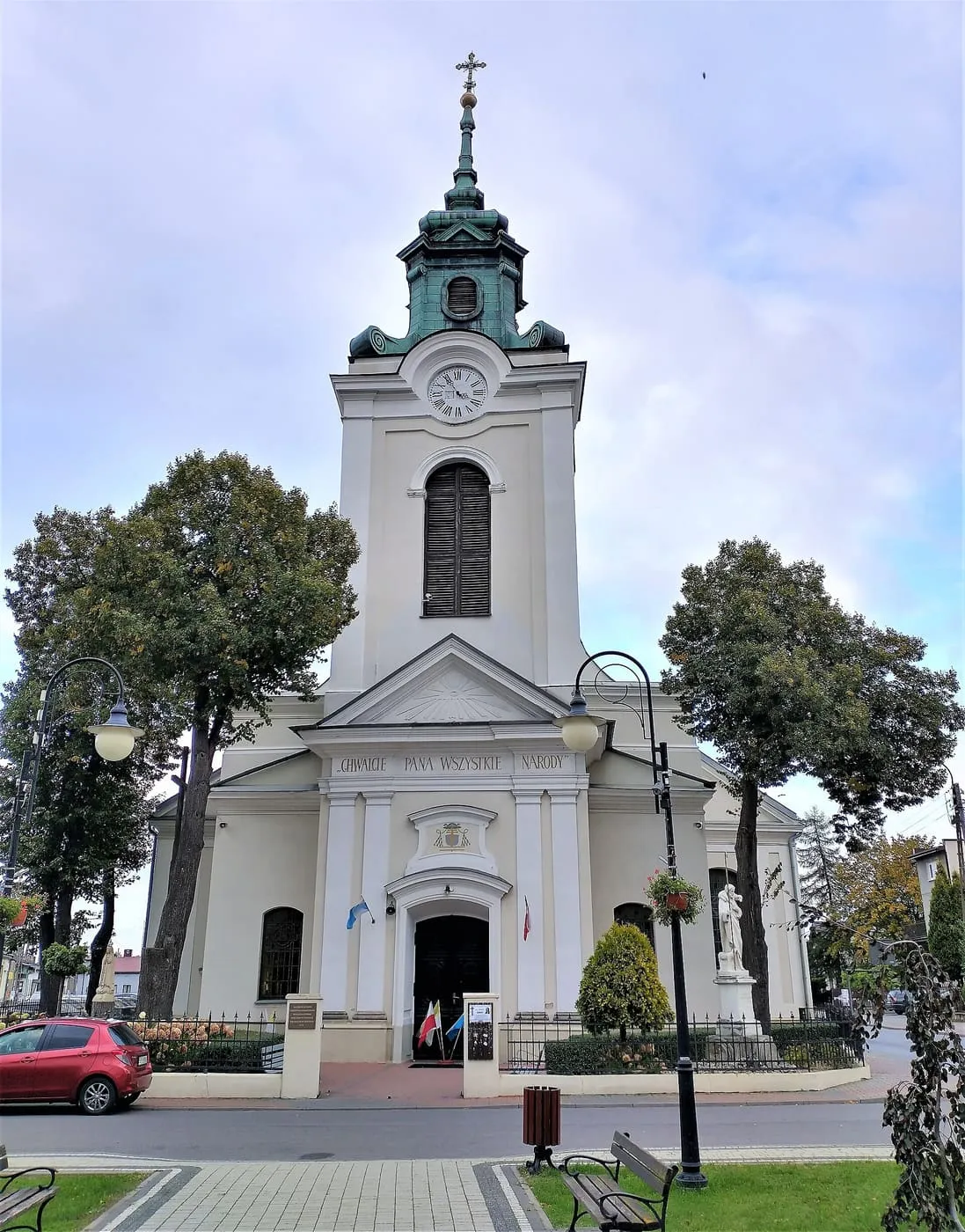 Biała fasada kolegiaty z wieżą z zegarem i zieloną, miedzianą kopułą z latarnią, otoczona drzewami; przed nią latarnie uliczne, ławka i chodnik