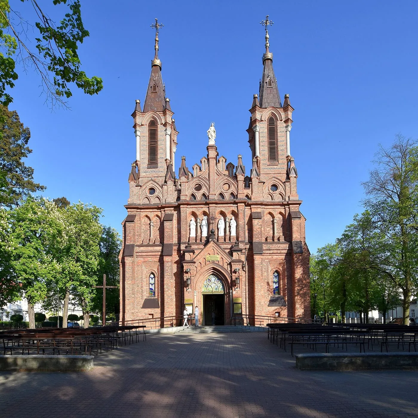 Ceglana, neogotycka fasada kolegiaty z dwiema smukłymi, ostro zakończonymi wieżami i bogatym portalem, przed nią brukowany plac z rzędami ławek