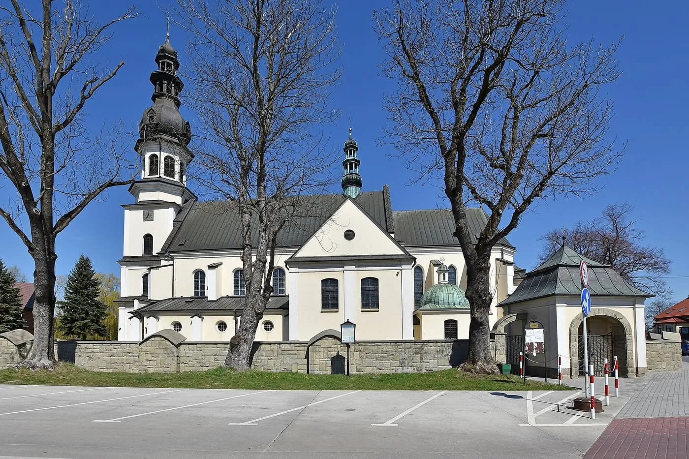 Biały kościół z ciemną, wielokondygnacyjną wieżą zwieńczoną cebulastym hełmem, obok murowana brama z daszkiem i kamienny mur, wśród bezlistnych drzew, na tle błękitnego nieba