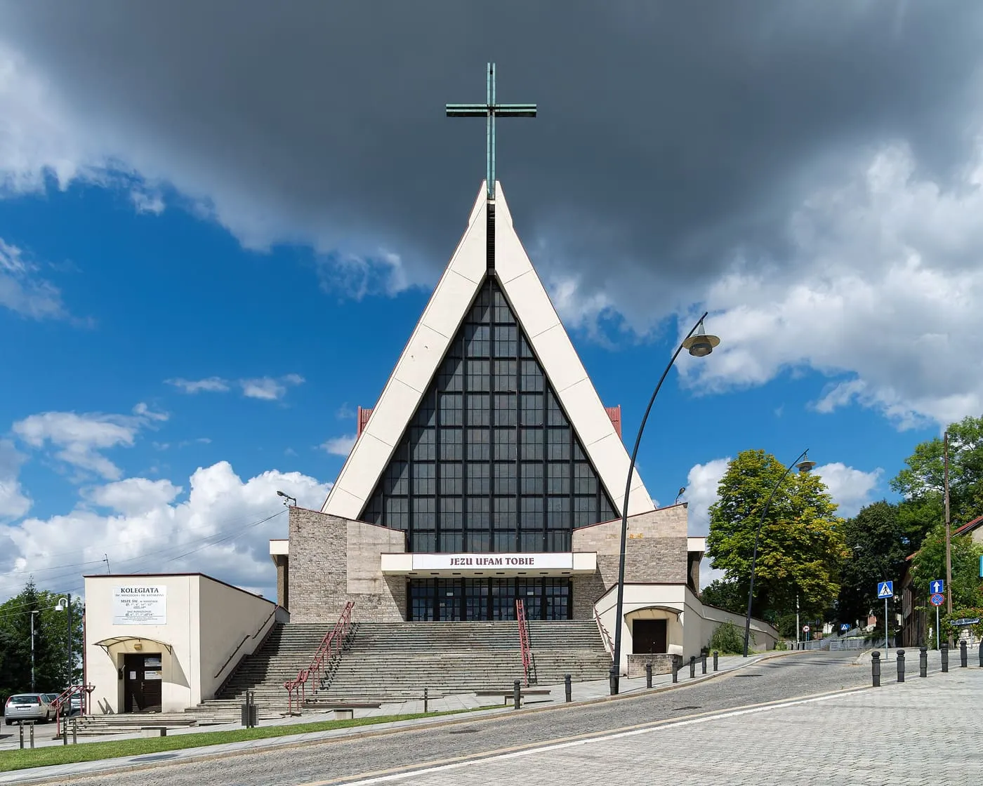 Nowoczesna fasada kolegiaty w kształcie trójkąta, ze szklaną ścianą i wysokim krzyżem, z szerokimi schodami przed wejściem i napisem „Jezu ufam Tobie”