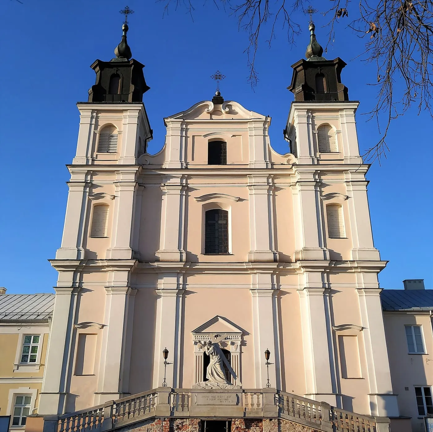 Późnobarokowa, jasnoróżowa fasada kolegiaty w Łukowie z dwiema wieżami nakrytymi ciemnymi, cebulastymi hełmami i figurą Chrystusa dźwigającego krzyż nad schodami z balustradą