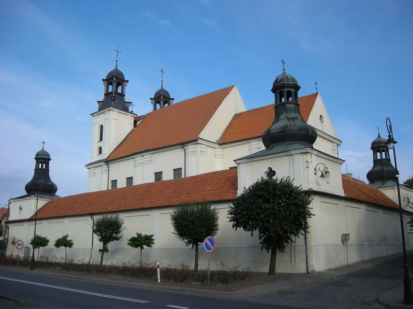Biało-żółta, barokowa kolegiata z dwiema wieżami zwieńczonymi ciemnymi hełmami i czerwonym dachem, widziana z ulicy za rzędem młodych drzew, na tle błękitnego nieba