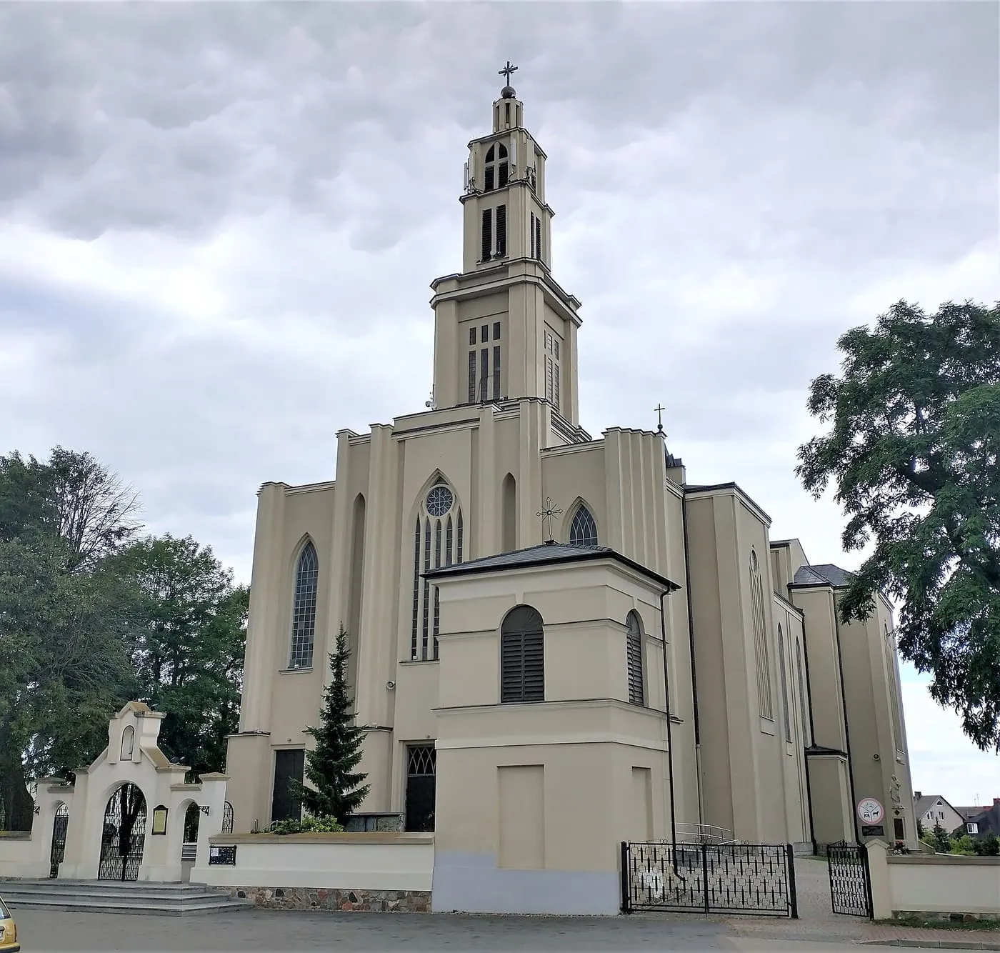 Jasna, modernistyczna fasada konkatedry z wysoką, smukłą wieżą i wąskimi oknami, za kutym ogrodzeniem z białą bramą, pod pochmurnym niebem