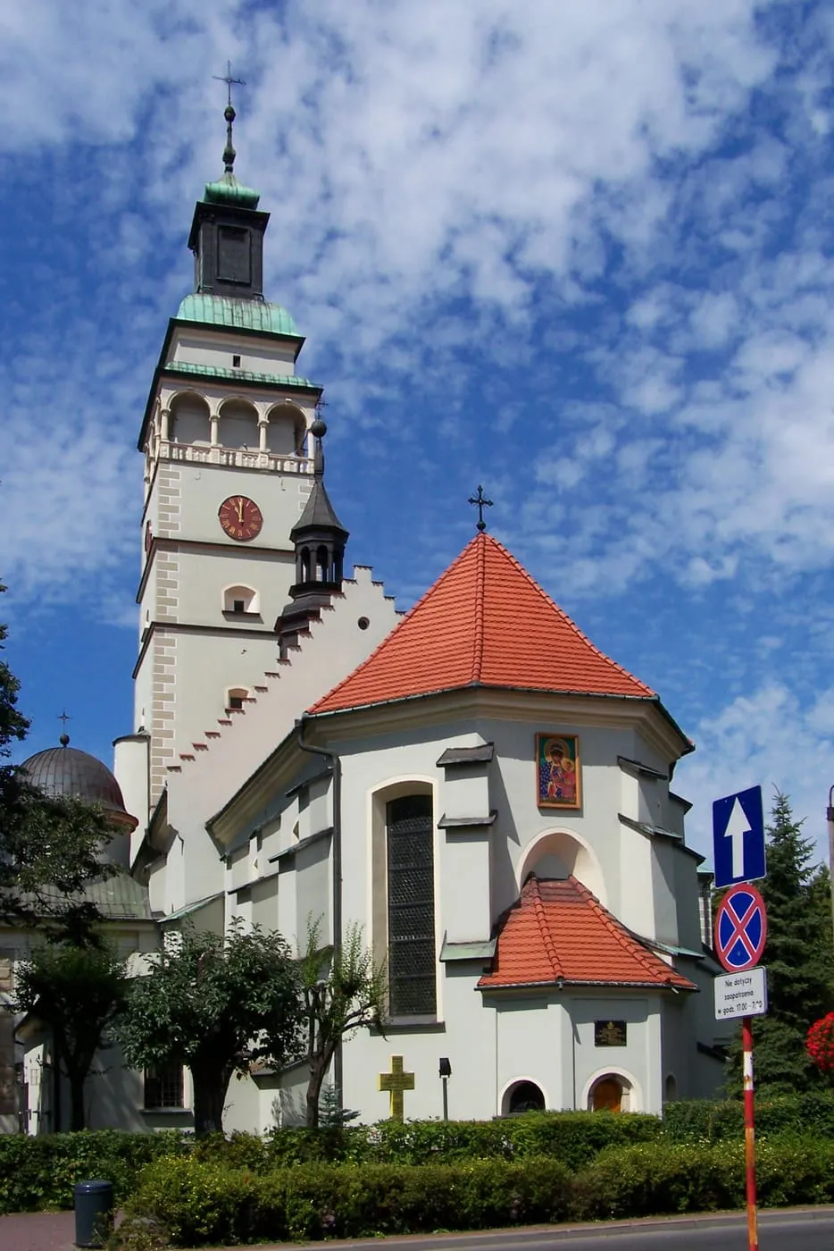 Wysoka, biała wieża konkatedry w Żywcu z balkonem i zegarem, obok czerwonego dachu kaplicy z ikoną Matki Bożej