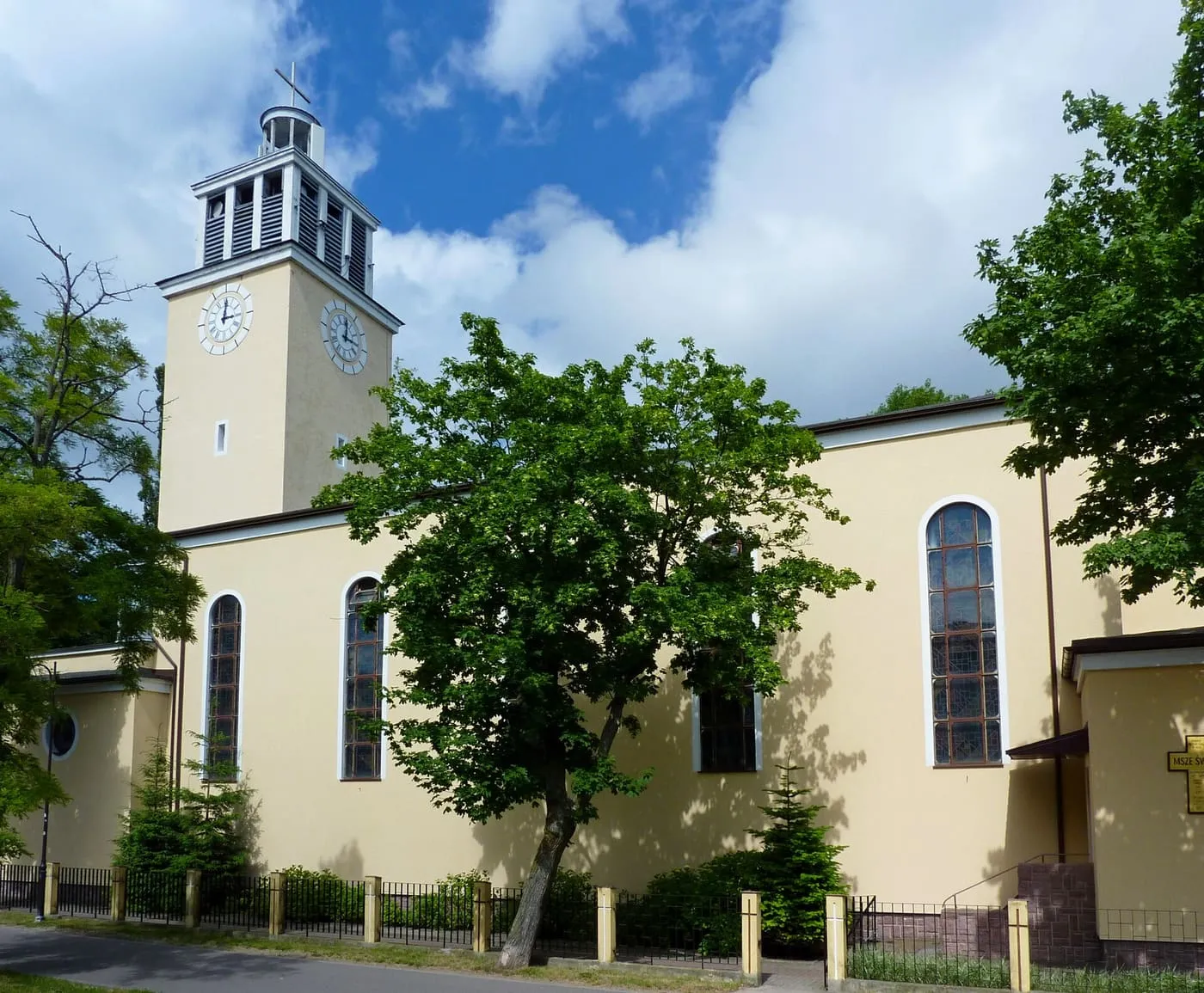 Żółtokremowy kościół z kwadratową wieżą z zegarem i otwartą latarnią dzwonną, z wysokimi łukowymi oknami nawy, za zielonymi drzewami i niskim ogrodzeniem