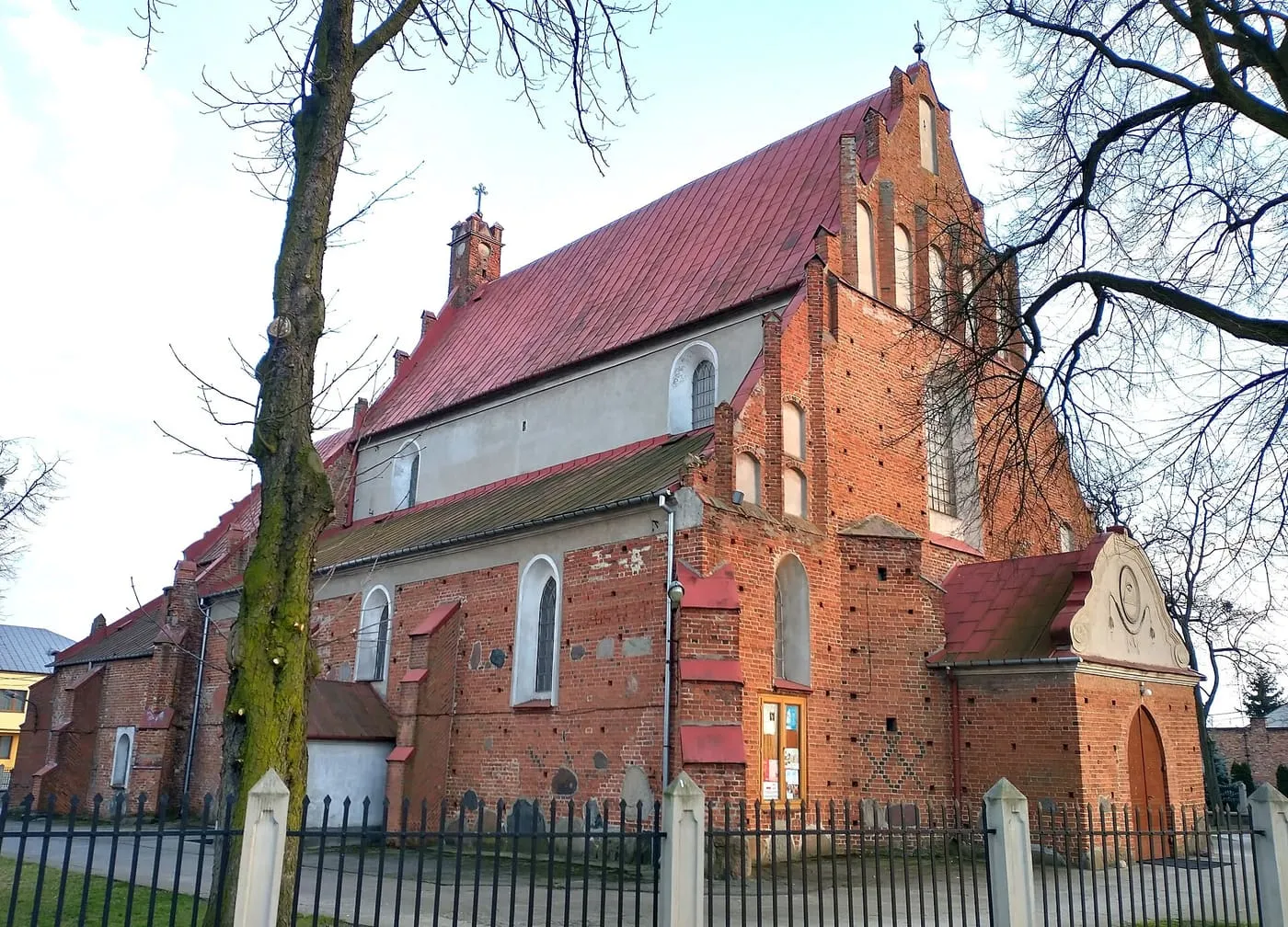 Gotycki kościół z czerwonej cegły, ze schodkowym szczytem i czerwonym, blaszanym dachem, widziany zza żelaznego ogrodzenia z jasnymi słupkami, wśród bezlistnych drzew