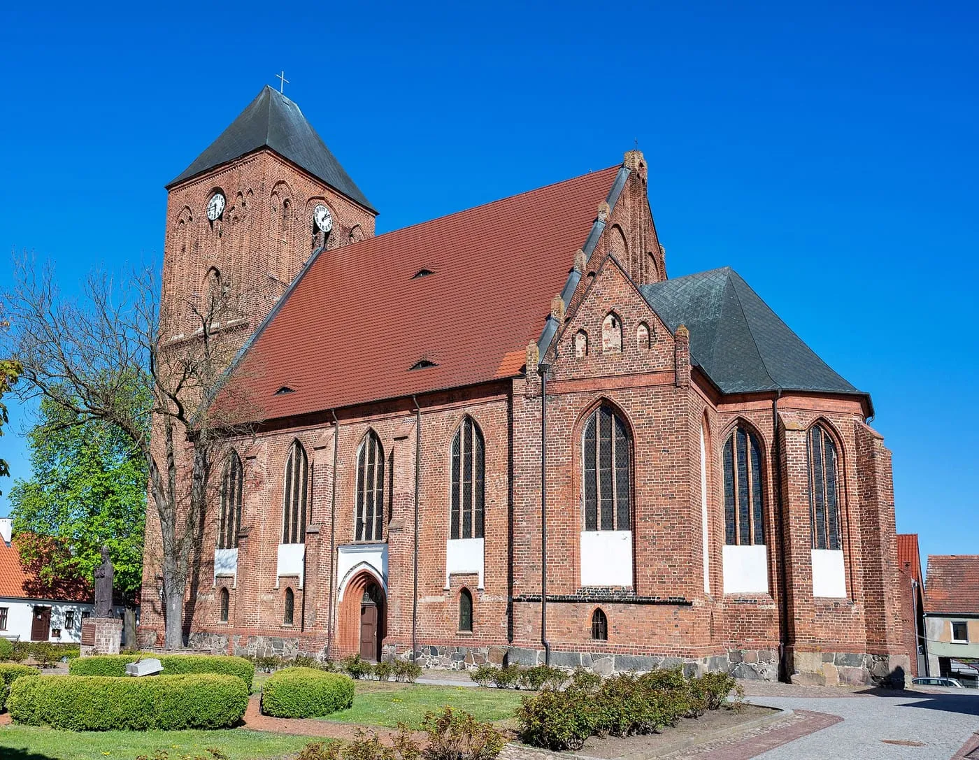 Gotycki, ceglany kościół z wysoką wieżą z zegarem i stromym czerwonym dachem, otoczony niskimi krzewami, na tle intensywnie błękitnego nieba