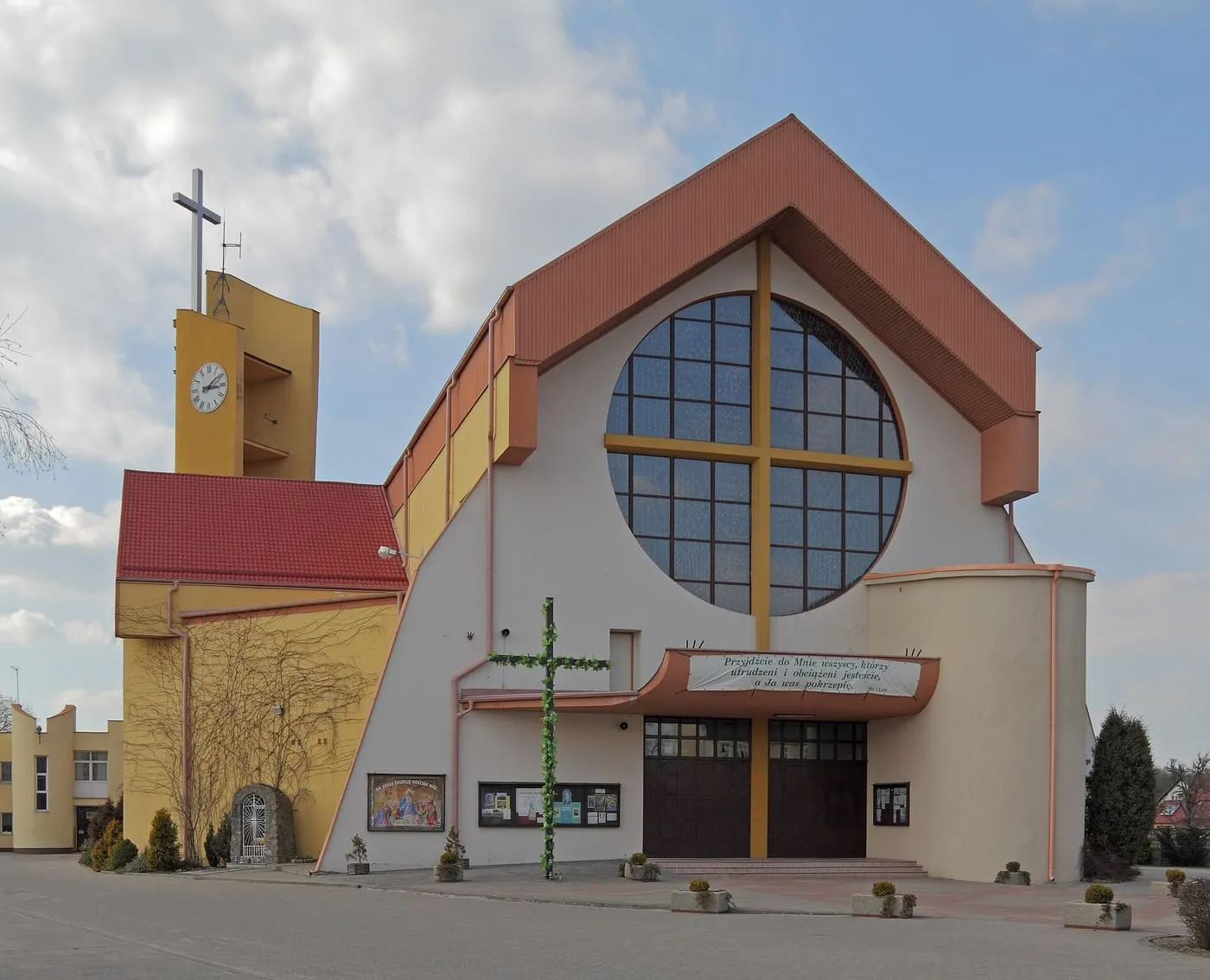 Nowoczesny kościół z wysokim, dwuspadowym dachem w kolorze miedzi, wielkim okrągłym oknem z krzyżem w fasadzie i żółtą wieżą z zegarem