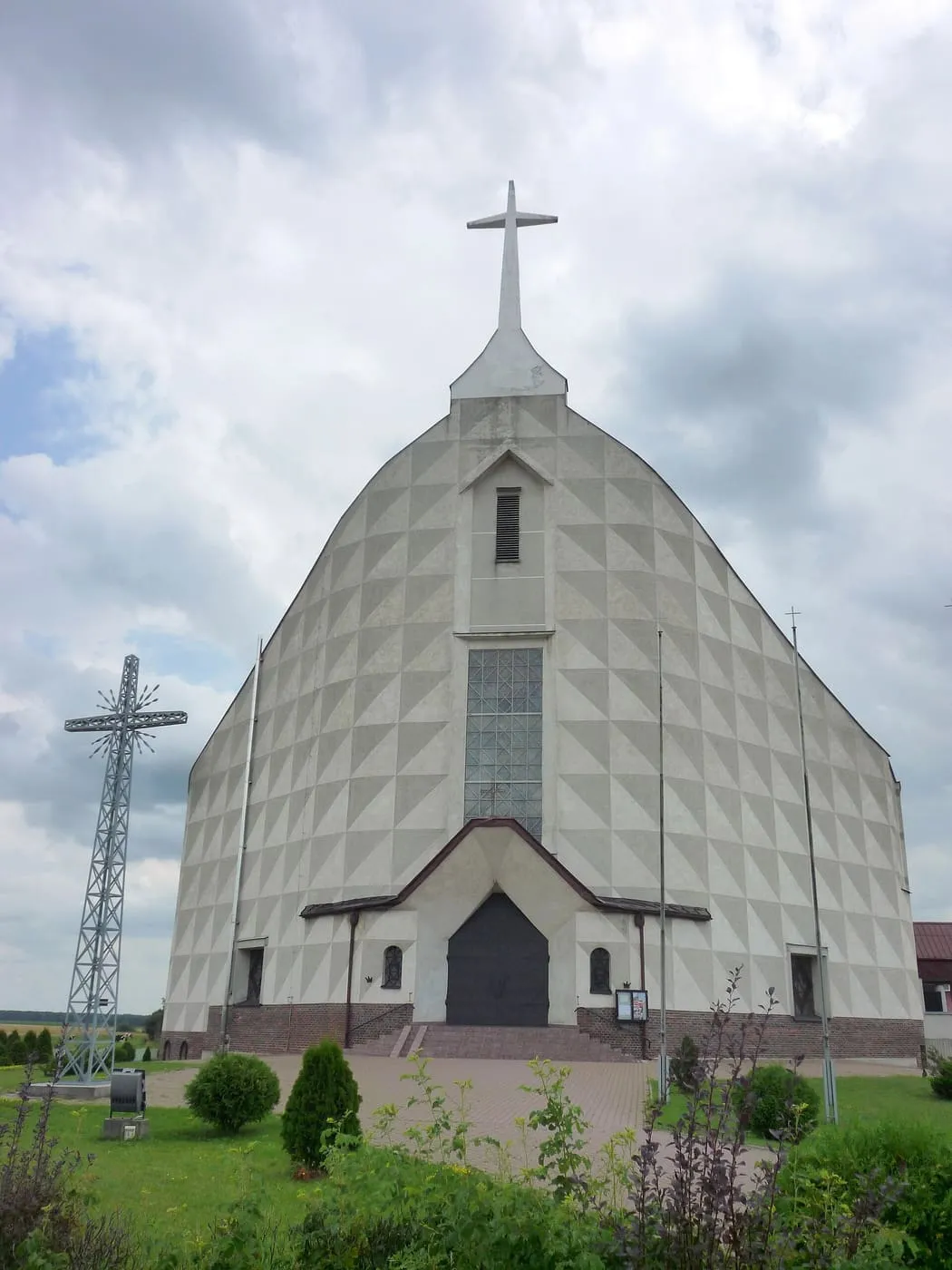 Nowoczesny kościół w kształcie trójkątnego namiotu, o ścianach z jasnych, geometrycznych płyt i z wysokim krzyżem na szczycie, przed nim trawnik i wysoki stalowy krzyż