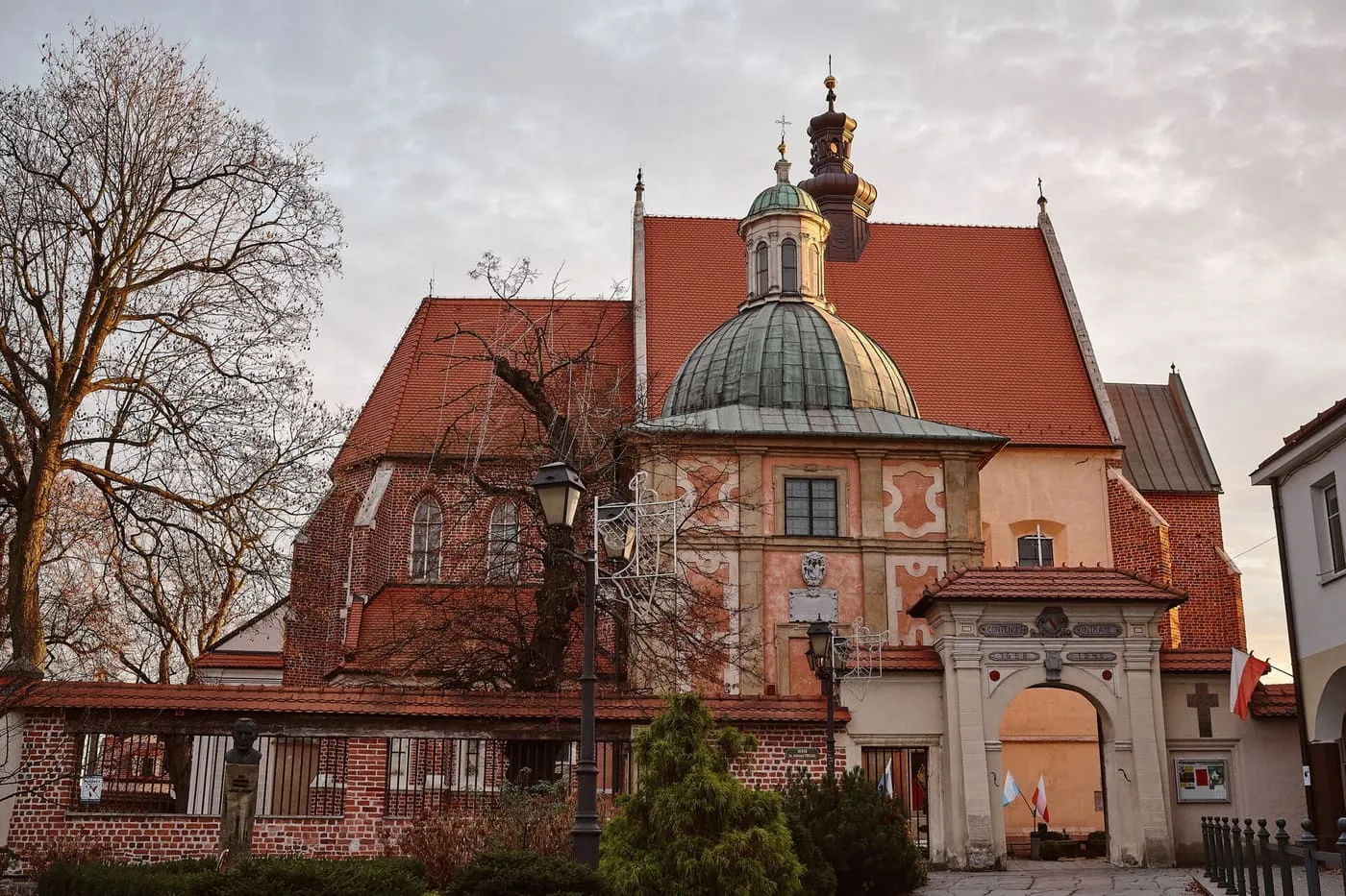 Gotycki kościół z czerwonym, stromym dachem i barokową, kopułową kaplicą z ozdobną fasadą, obok brama, wśród drzew w świetle zachodzącego słońca