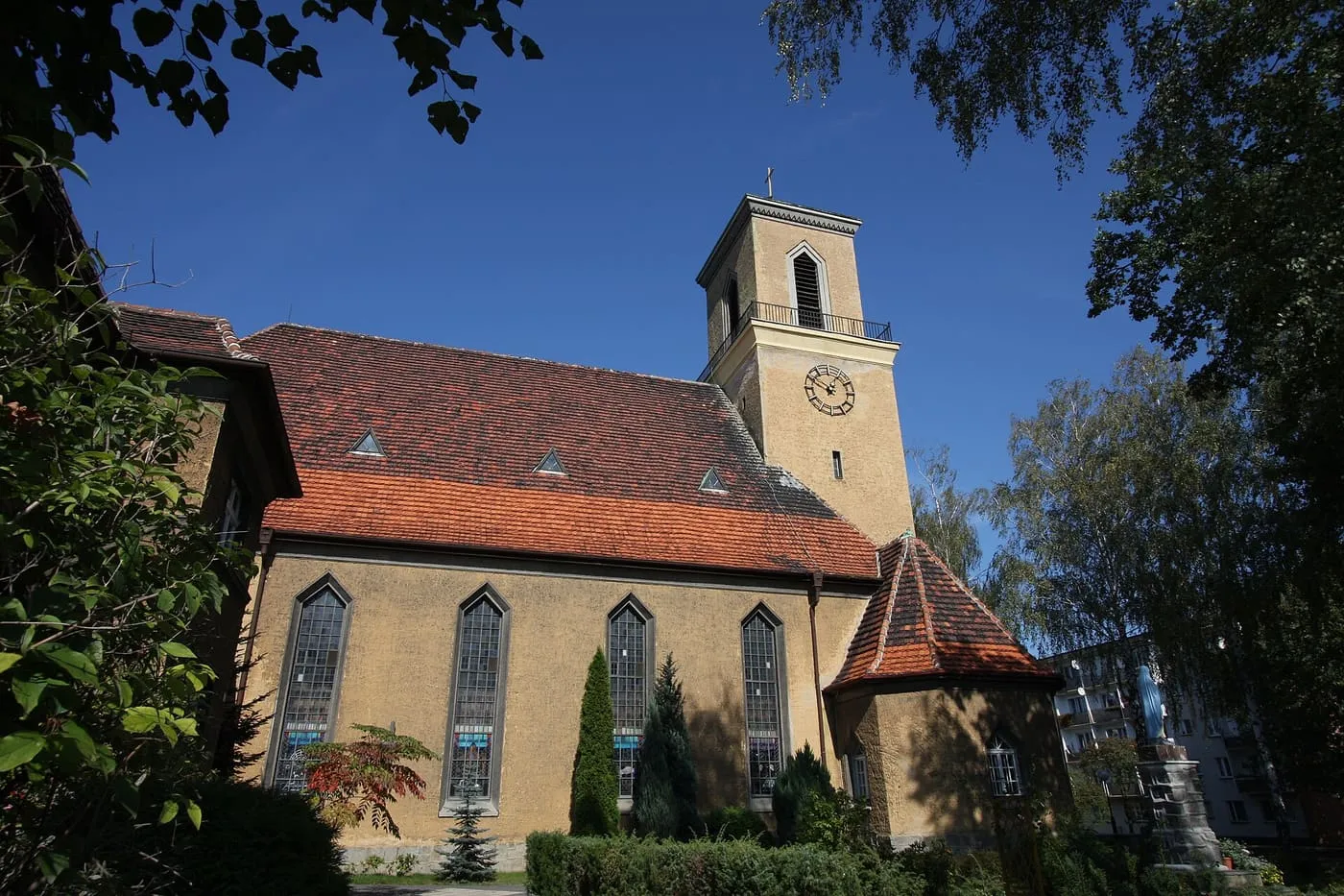 Żółty kościół z cegły klinkierowej z wysokimi, wąskimi oknami, czerwonym dachem i kwadratową wieżą z zegarem, widziany spod jesiennych drzew na tle błękitnego nieba