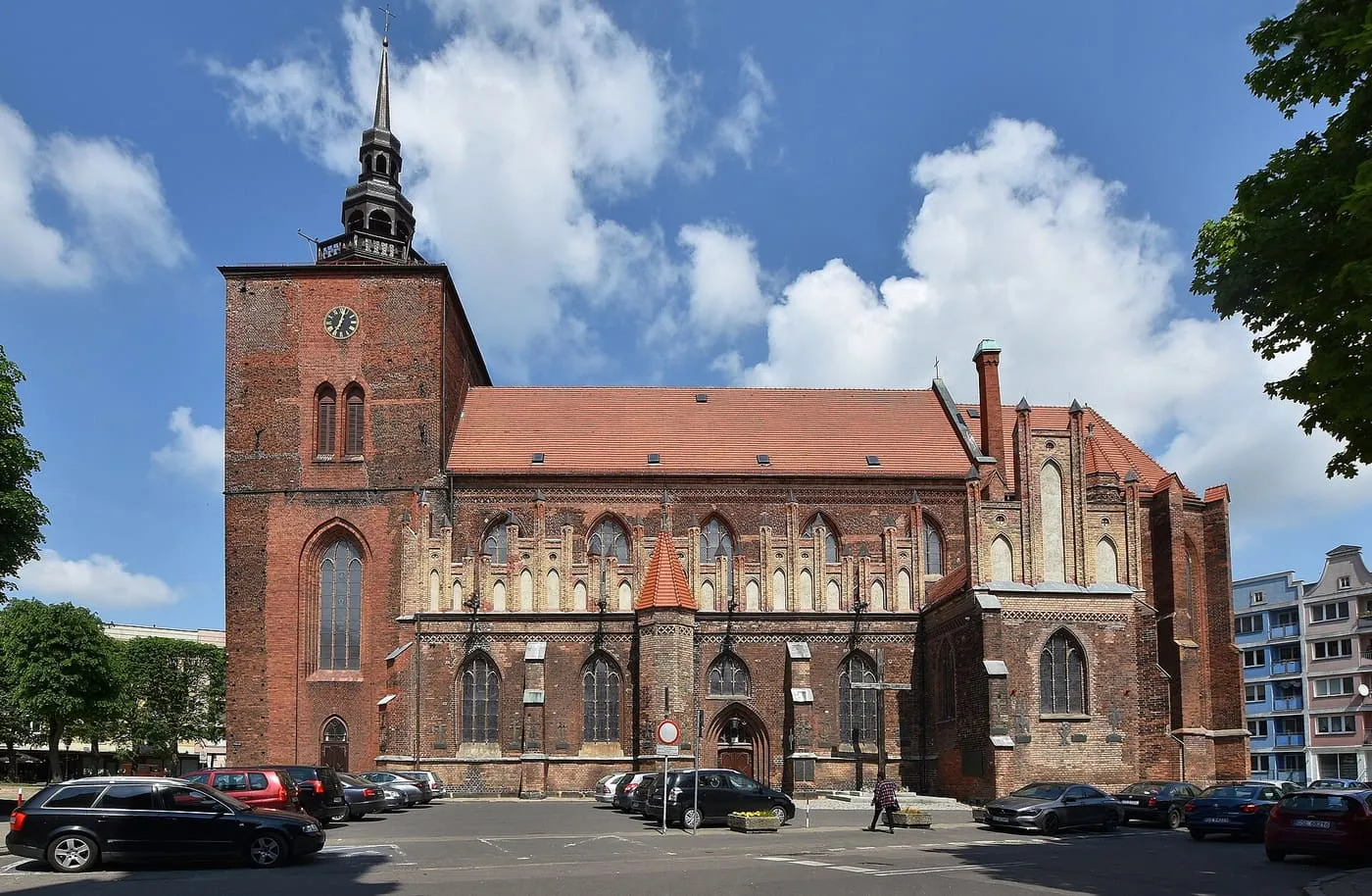 Ceglany, gotycki kościół Mariacki w Słupsku z masywną wieżą z zegarem i smukłą iglicą oraz długim, czerwonym dachem nad korpusem, widziany z parkingu