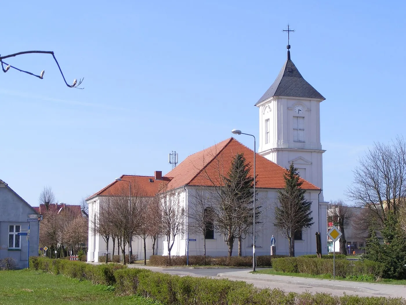 Biały kościół z pomarańczowym dachem i wieżą z ciemnym, stożkowym hełmem, obok skwer i ulica