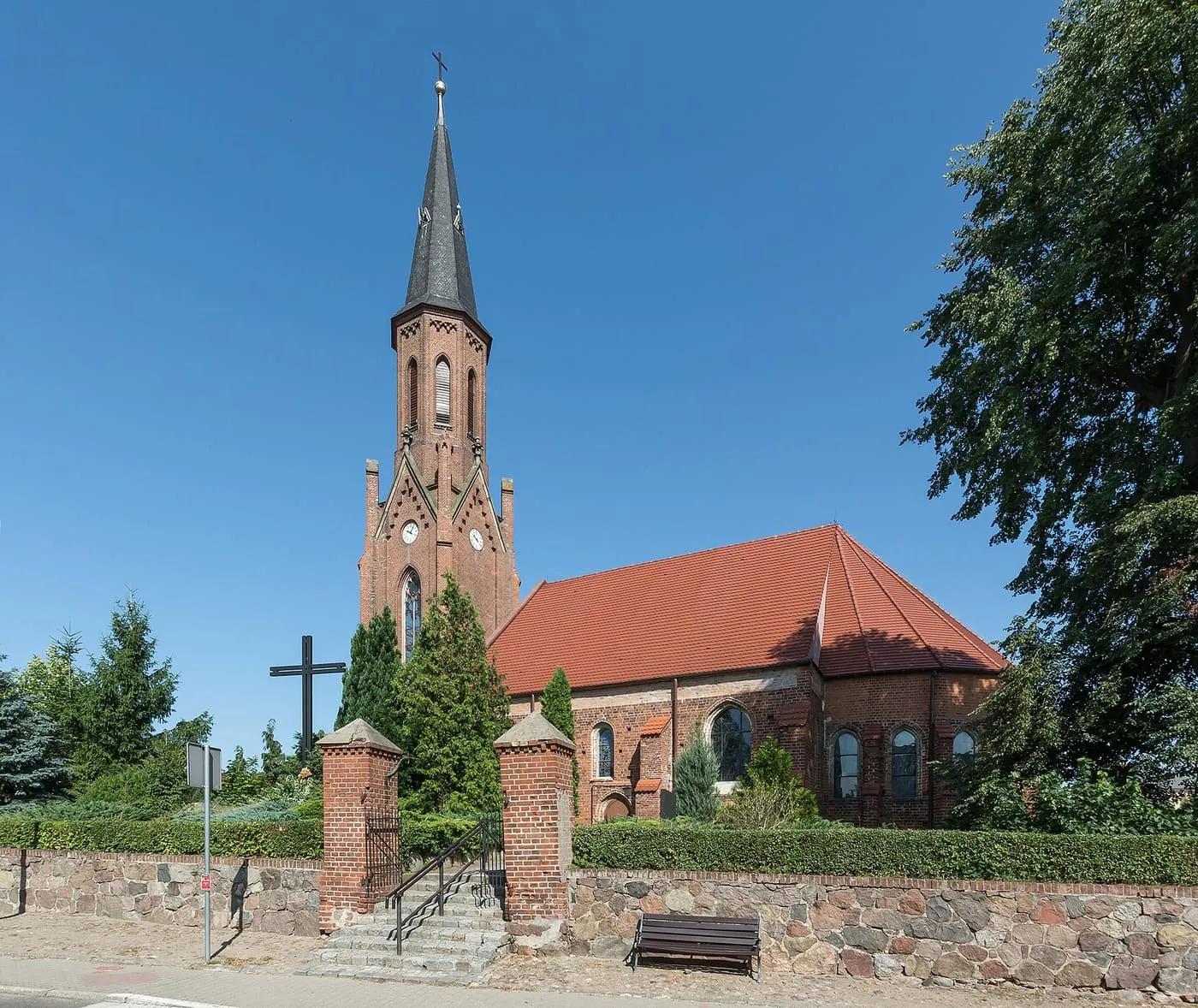 Ceglany kościół z wysoką, neogotycką wieżą i czerwonym dachem, otoczony kamiennym murem i drzewami, z krzyżem przed wejściem, na tle błękitnego nieba