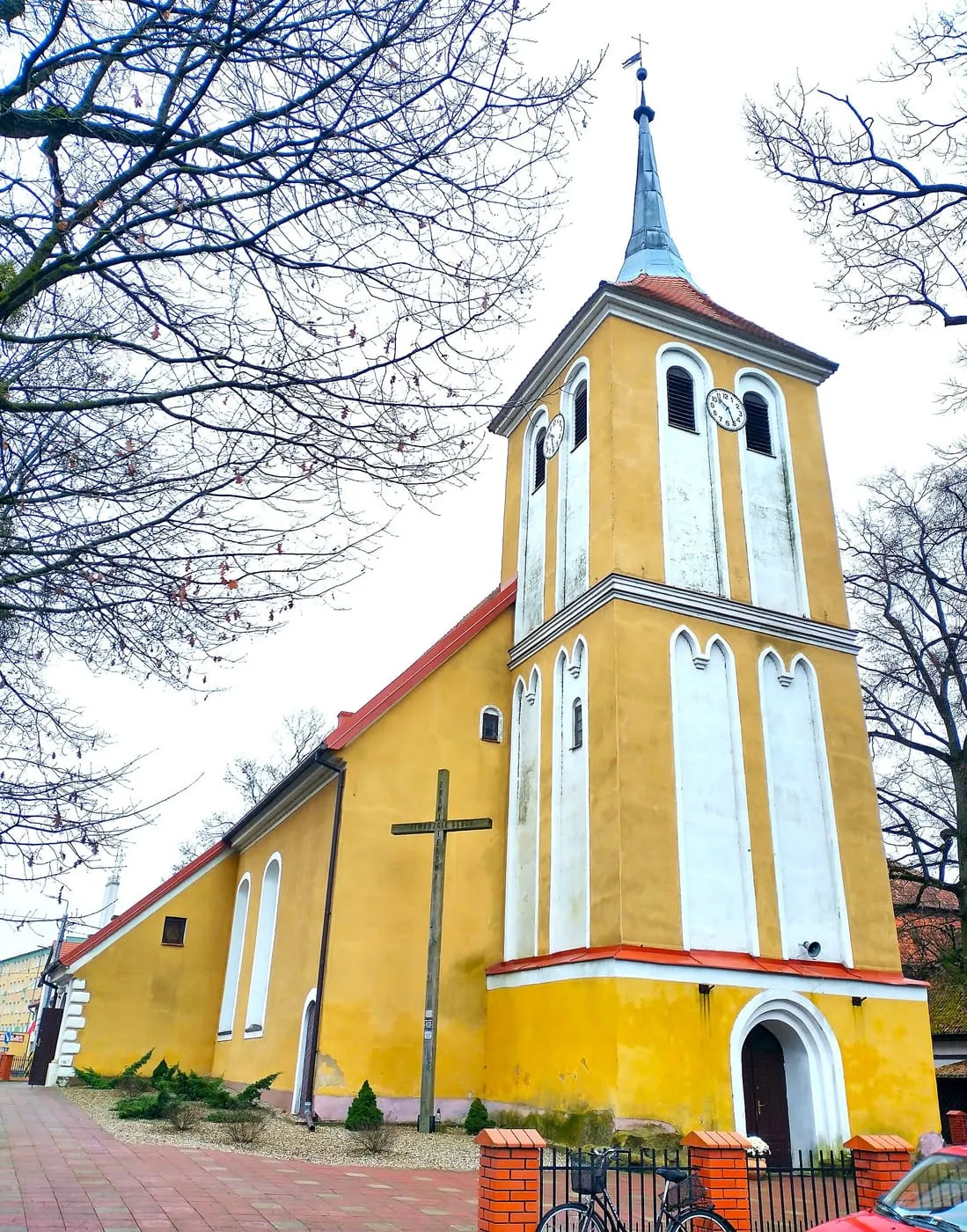 Żółty kościół z białymi pilastrami i wysoką wieżą z zegarem i niebieskawym hełmem, przy kościele duży krzyż, wśród gołych drzew