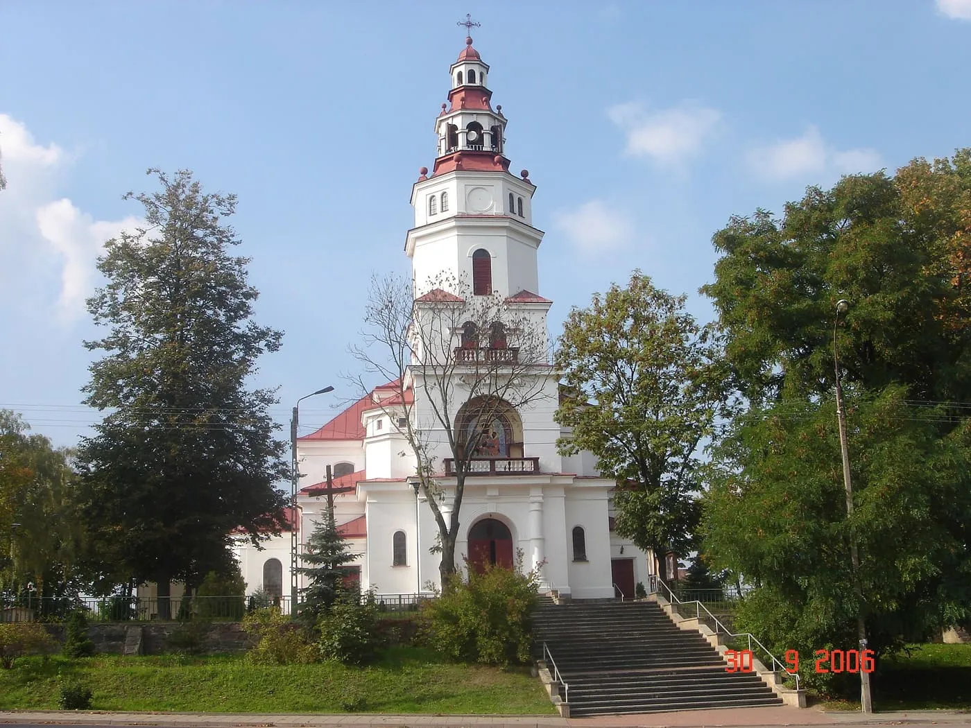 Biały kościół z wysoką, smukłą wieżą zwieńczoną czerwonym hełmem z latarnią, do którego prowadzą szerokie schody; wokół rosną wysokie drzewa