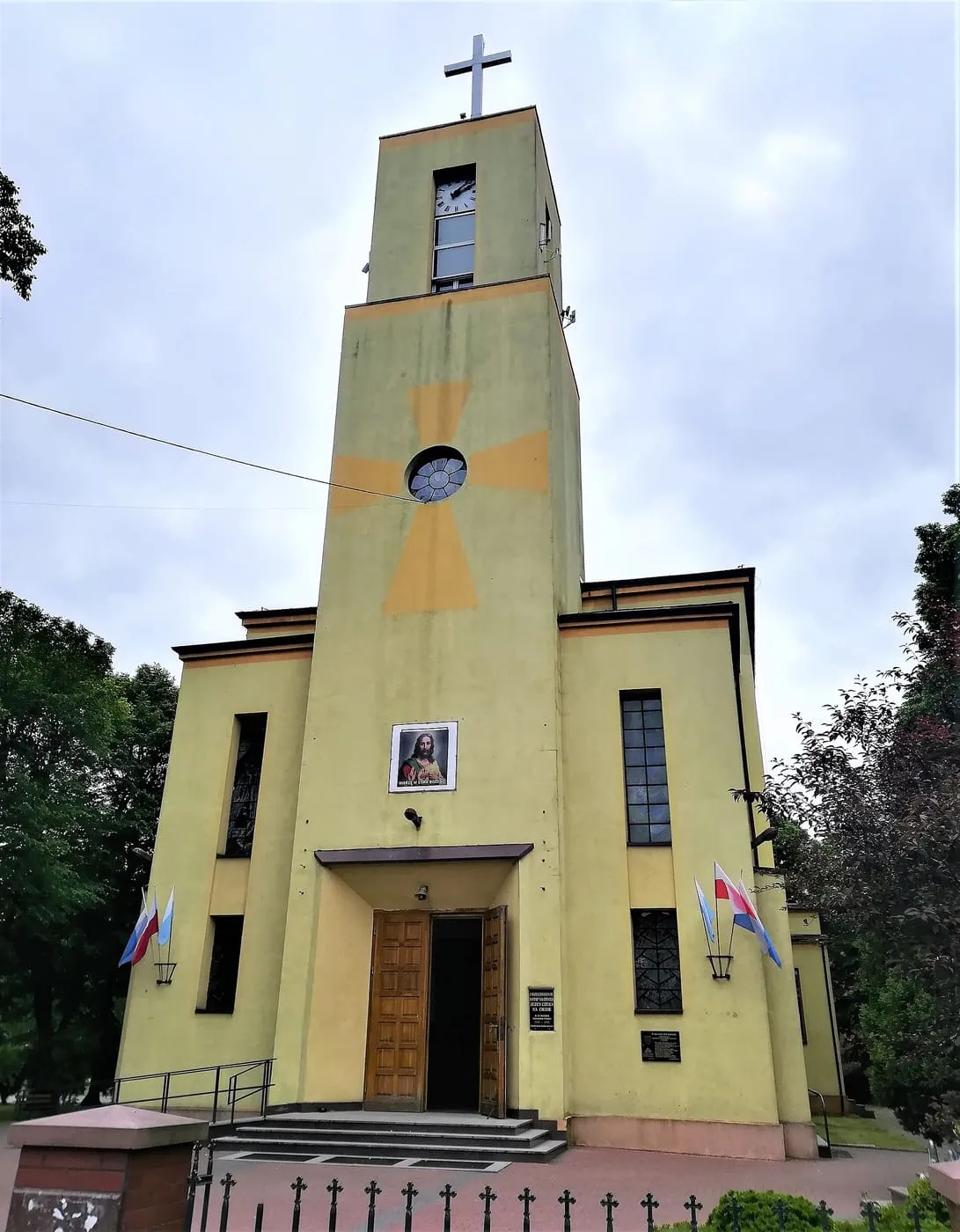 Modernistyczny kościół z prostą, wysoką wieżą-fasadą w odcieniu żółci, z krzyżem na szczycie, przed którym stoją schody i ogrodzenie