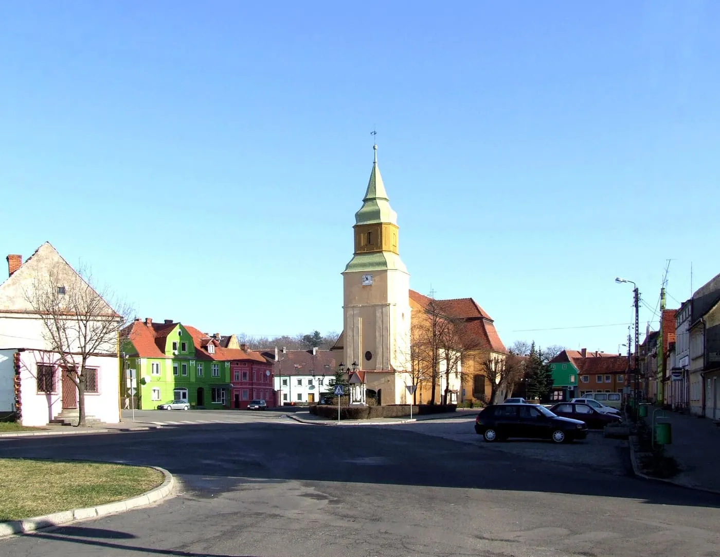Rynek z kolorowymi kamienicami, pośrodku którego stoi żółto-zielona wieża kościoła z ostrym hełmem, a obok pomarańczowy dach nawy; na jezdni zaparkowane samochody