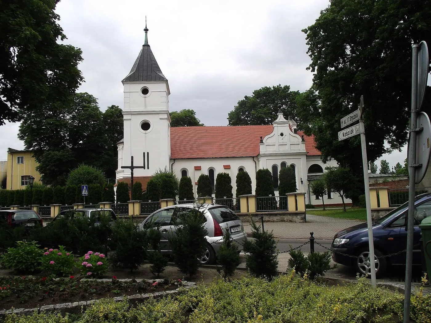 Biały kościół z czworoboczną wieżą z ciemnym dachem i czerwonym dachem nawy, za żywopłotem i kwiatowymi rabatami, z zaparkowanymi samochodami i tablicą z nazwami ulic