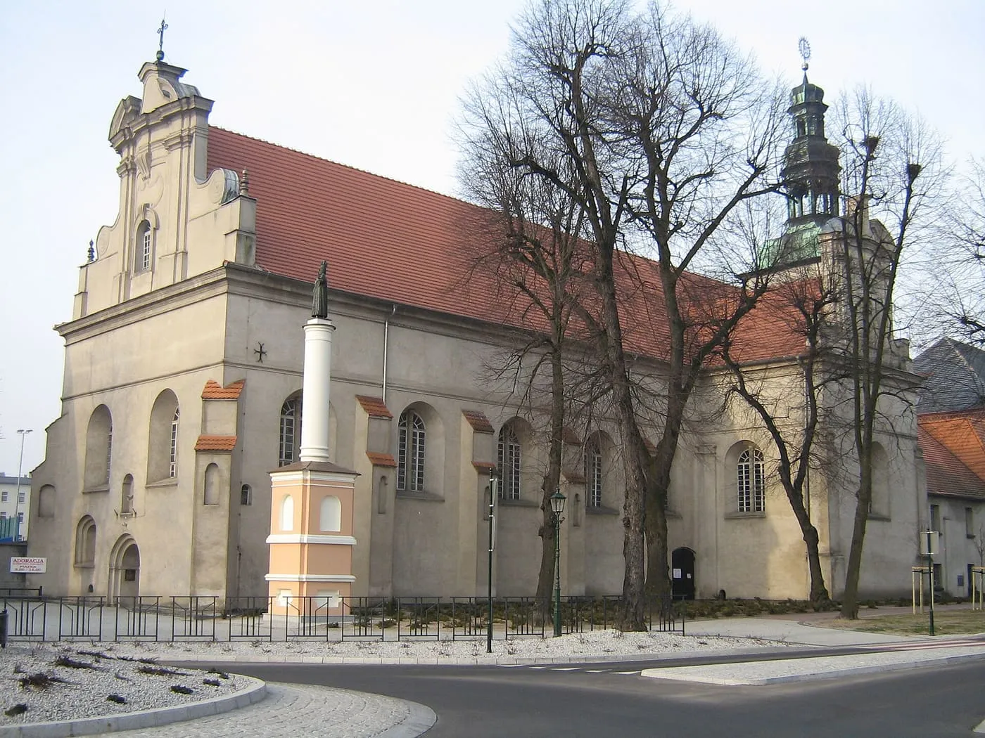 Kremowa, barokowa fasada kościoła pobernardyńskiego w Grodzisku Wielkopolskim z falistym szczytem i figurą Matki Boskiej na kolumnie przed wejściem, z wieżą klasztorną widoczną w tle