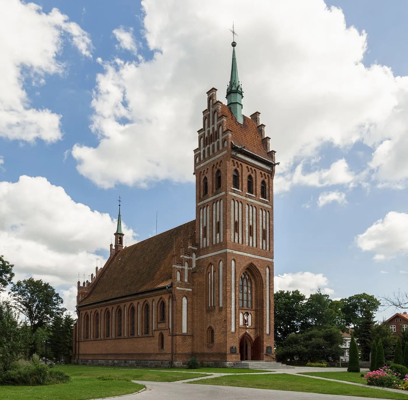Ceglany, neogotycki kościół z wysoką wieżą o schodkowych szczytach i zielonym hełmie, otoczony zielenią i chmurami